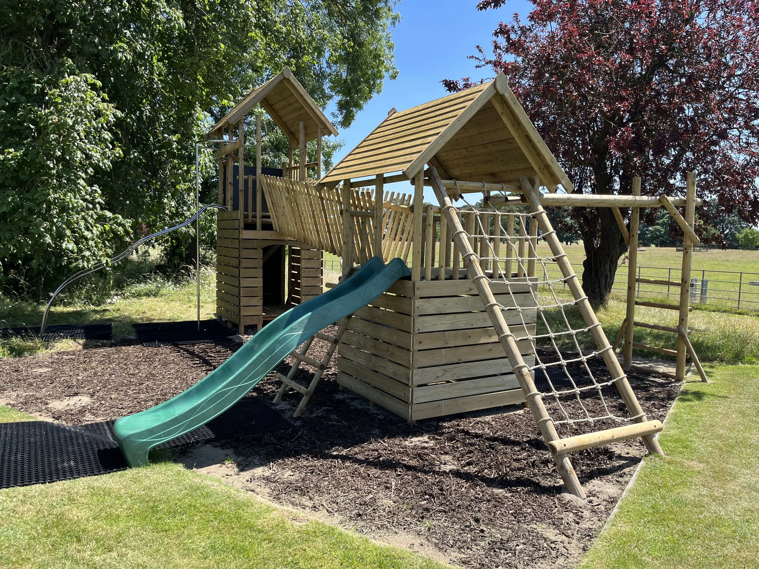 Wooden outdoor playground structure with slide, climbing ladders, and towers, surrounded by grass, trees, and fence.