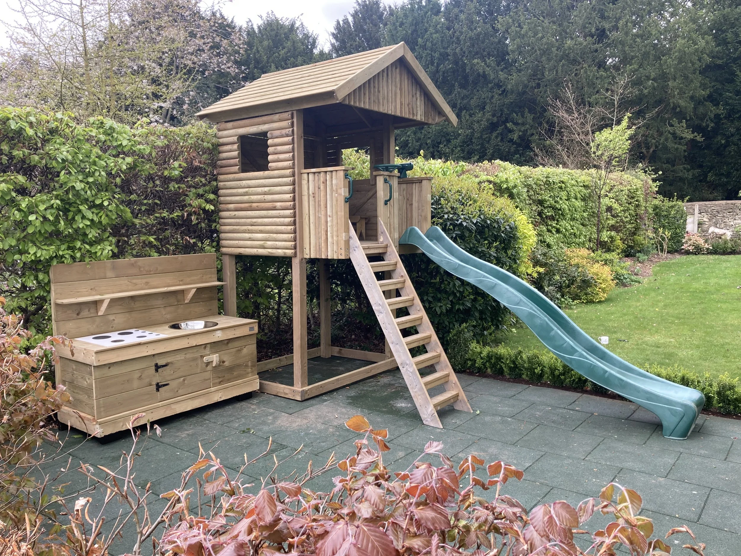 Children's wooden outdoor playset with slide and outdoor kitchen in a backyard with green bushes and trees.