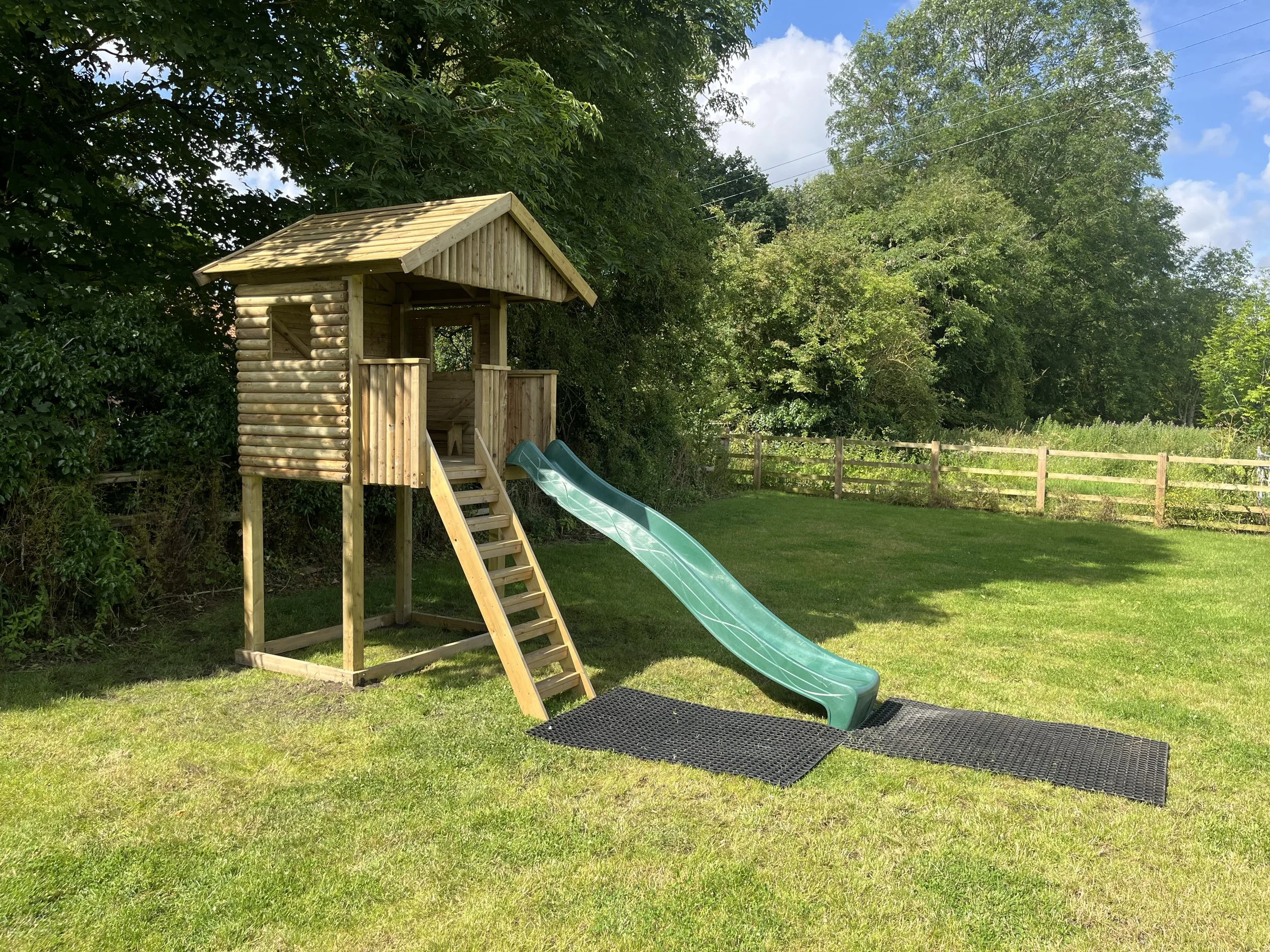 A wooden playhouse with a slide in a grassy backyard with trees and a wooden fence.