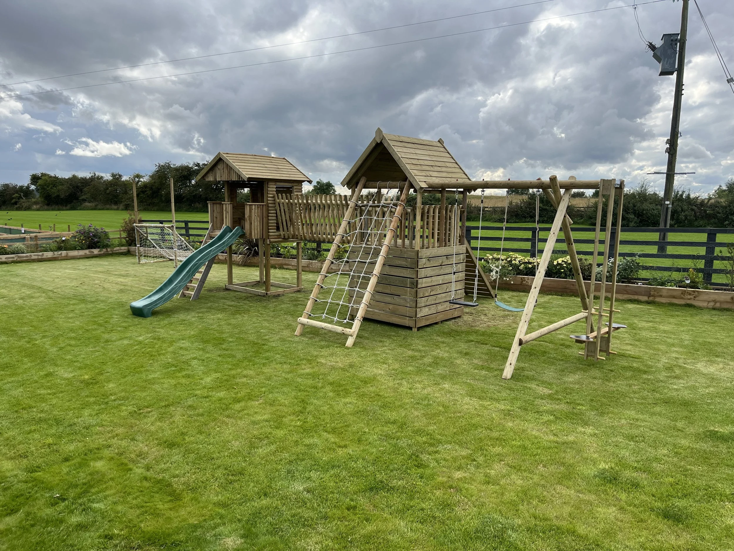 A wooden children’s playset with a slide, climbing ropes, swings, and a small house on a grassy yard under a cloudy sky.