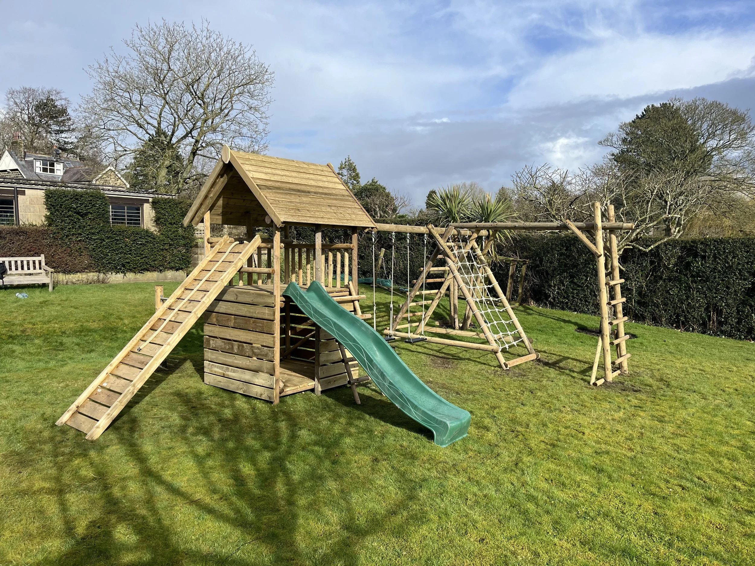 Wooden playground set with slide, climbing walls, swings, and ladder on a grassy yard, with trees and a hedge in the background under a partly cloudy sky.