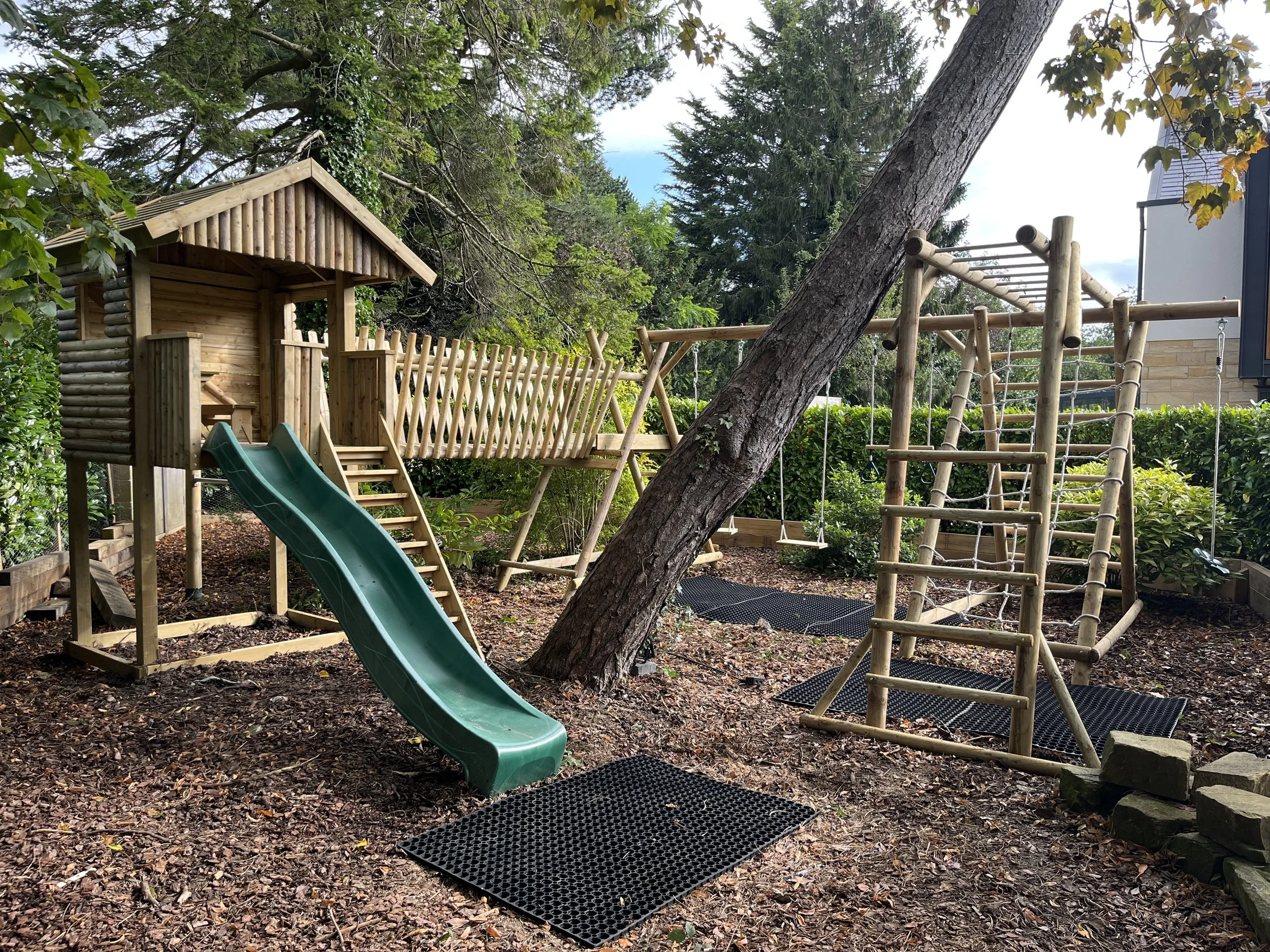 Children's outdoor wooden playset with a slide, swings, and climbing structures in a backyard with trees and bushes.