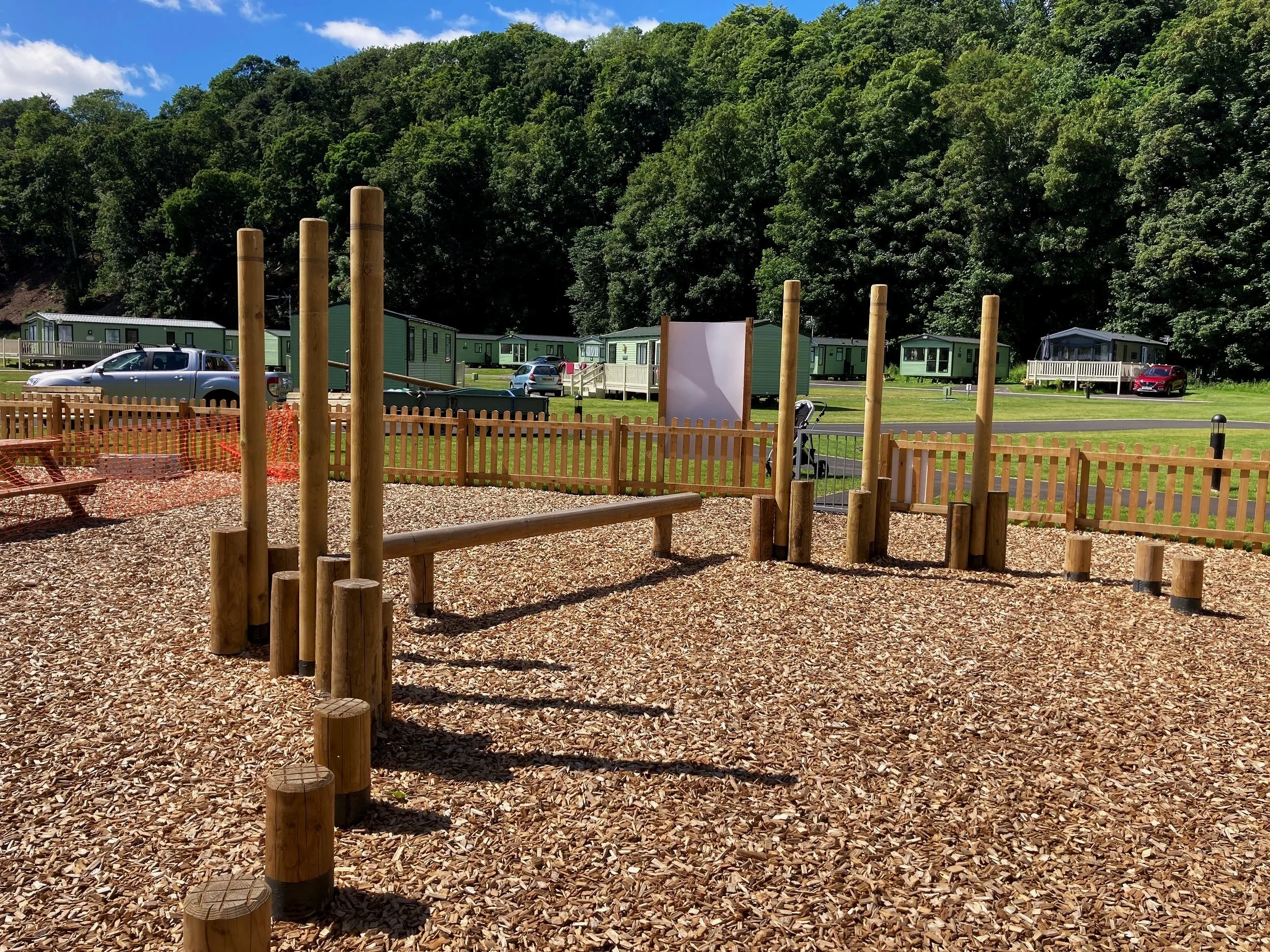 A playground with wooden climbing structures on wood chip ground, enclosed by a wooden fence, with mobile homes and cars parked in the background and green trees behind.
