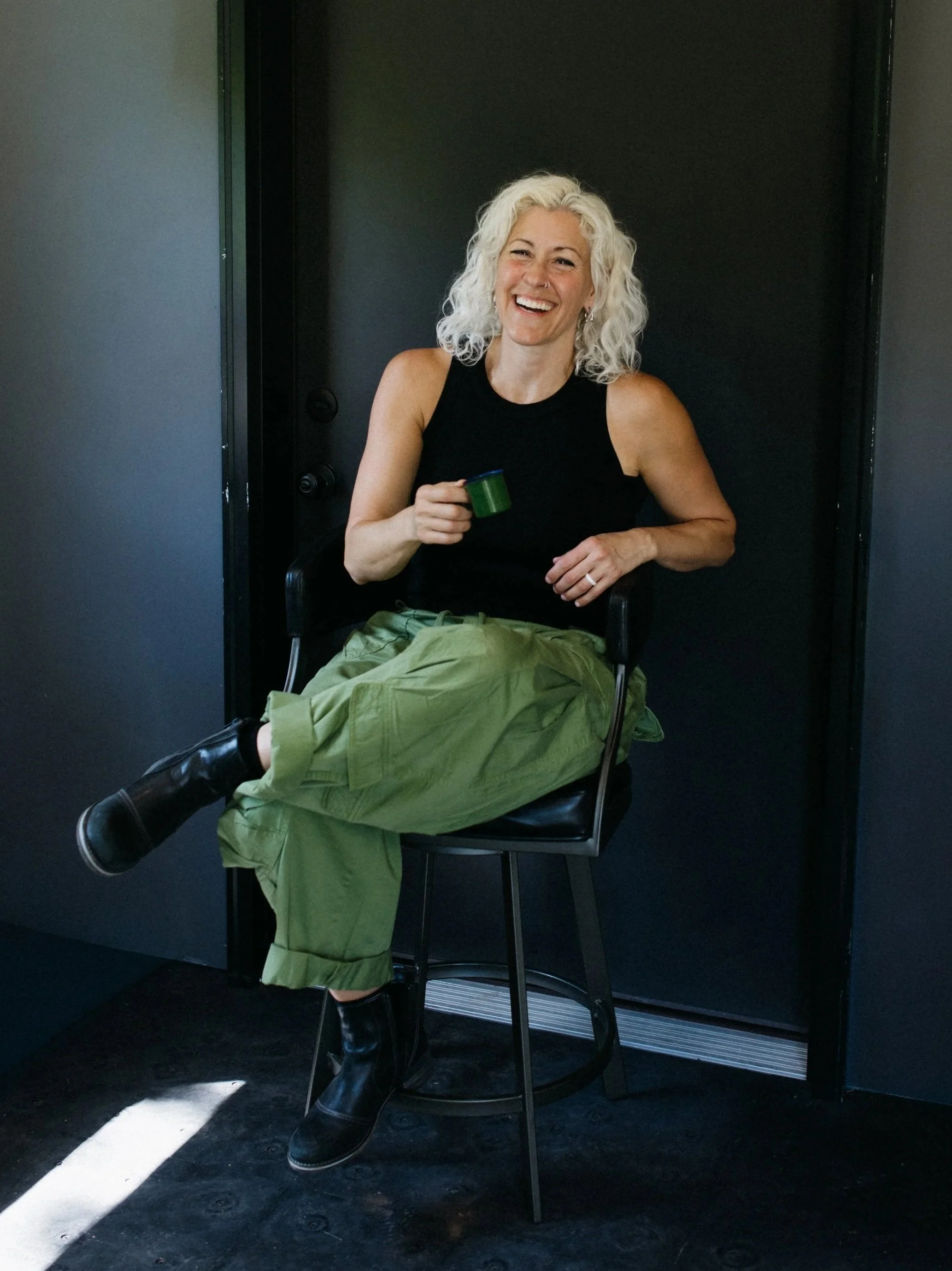 A woman with curly platinum blonde hair, sitting on a black chair and smiling, holding a green cup, wearing a black sleeveless top, olive green pants, and black boots, against a black backdrop.