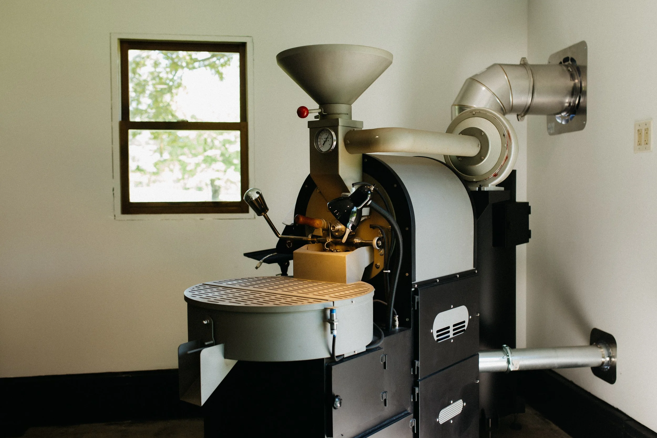 A coffee roaster machine in a room with a window showing trees outside.