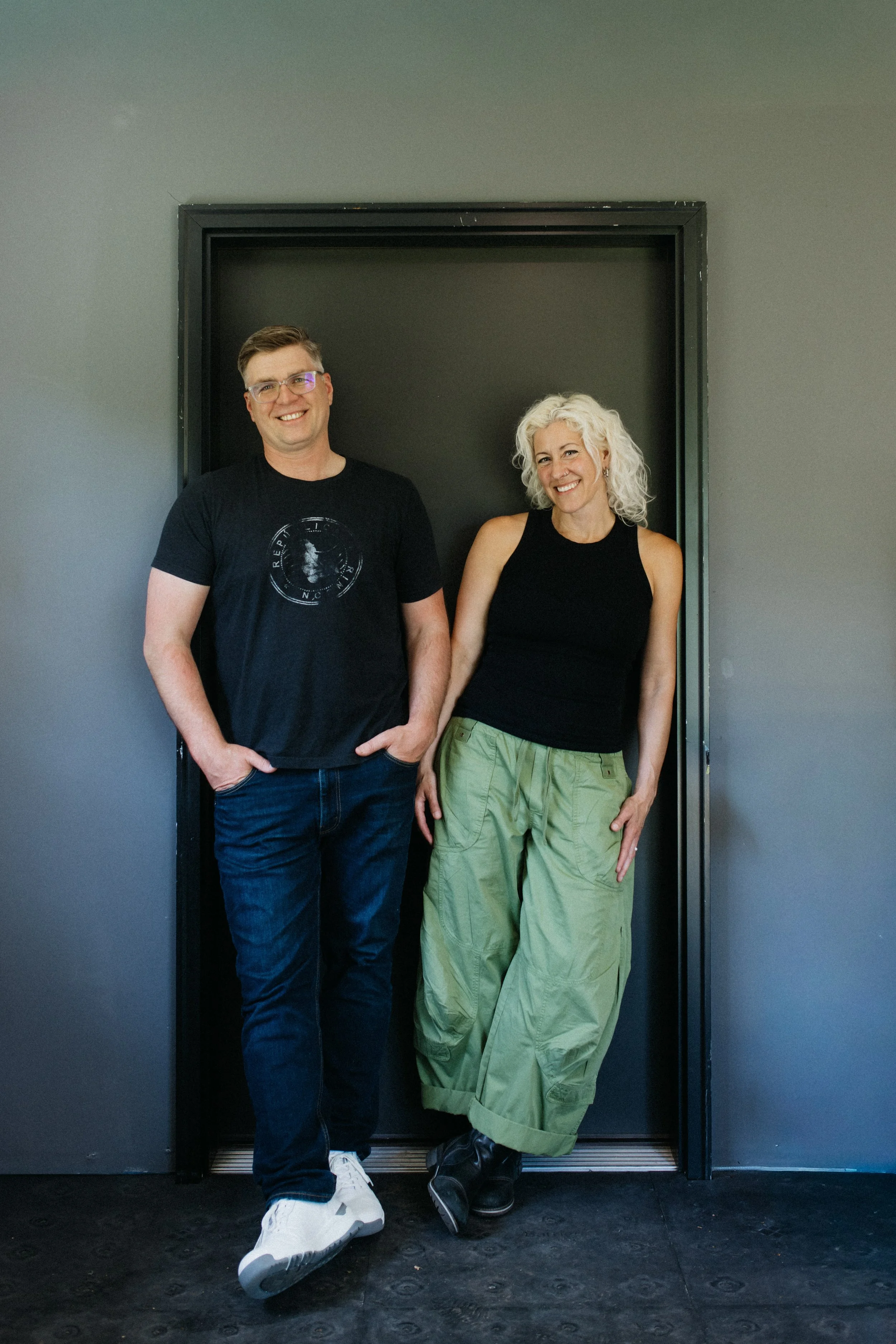 A man and a woman standing inside an elevator with black walls and a black floor, smiling at the camera. The man is wearing glasses, a black t-shirt, and jeans, and the woman is wearing a black sleeveless top and green cargo pants.