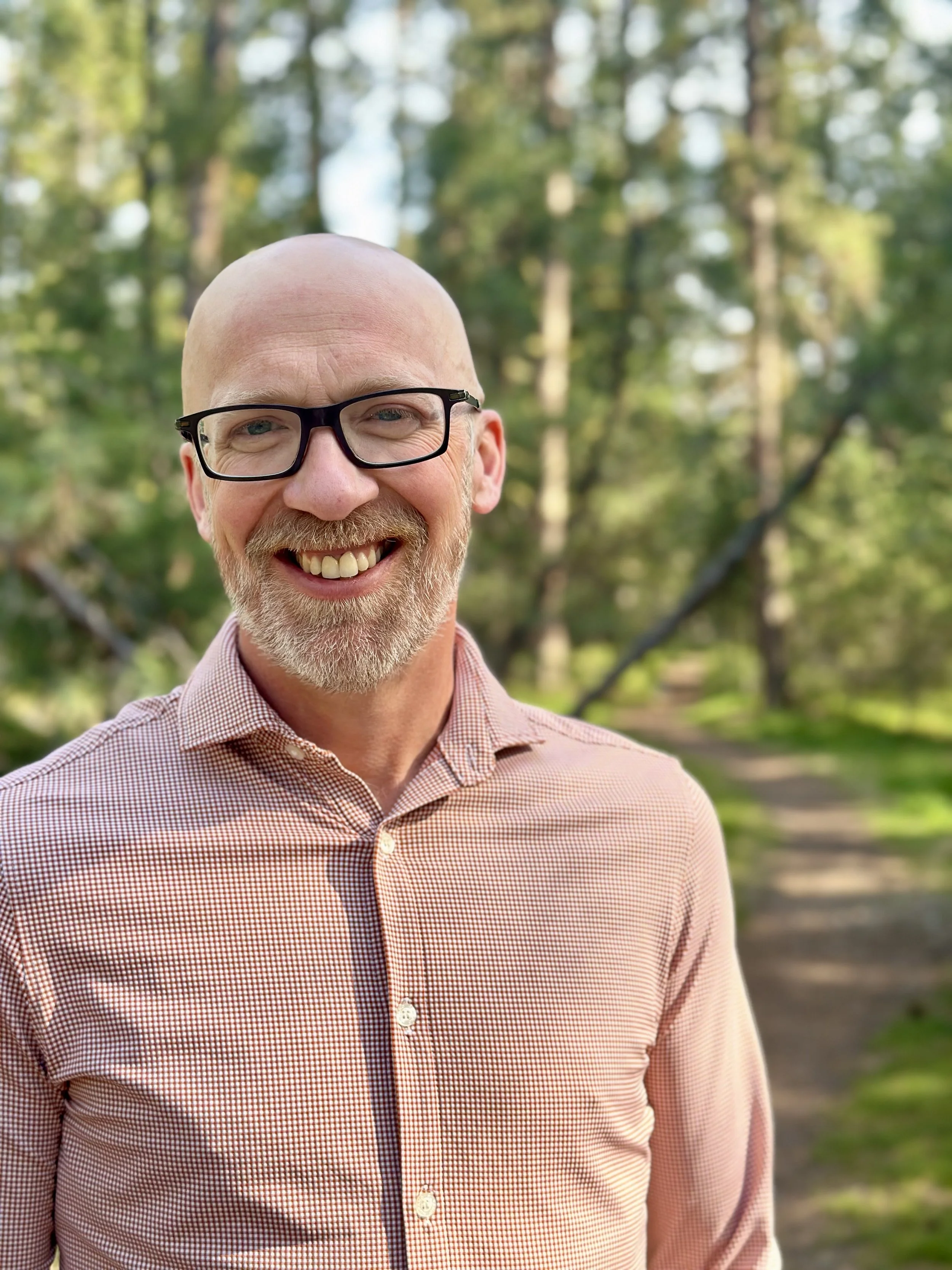 Close-up of smiling middle-aged man with glasses and a striped pink shirt outdoors in a park with trees and a walking trail in the background.