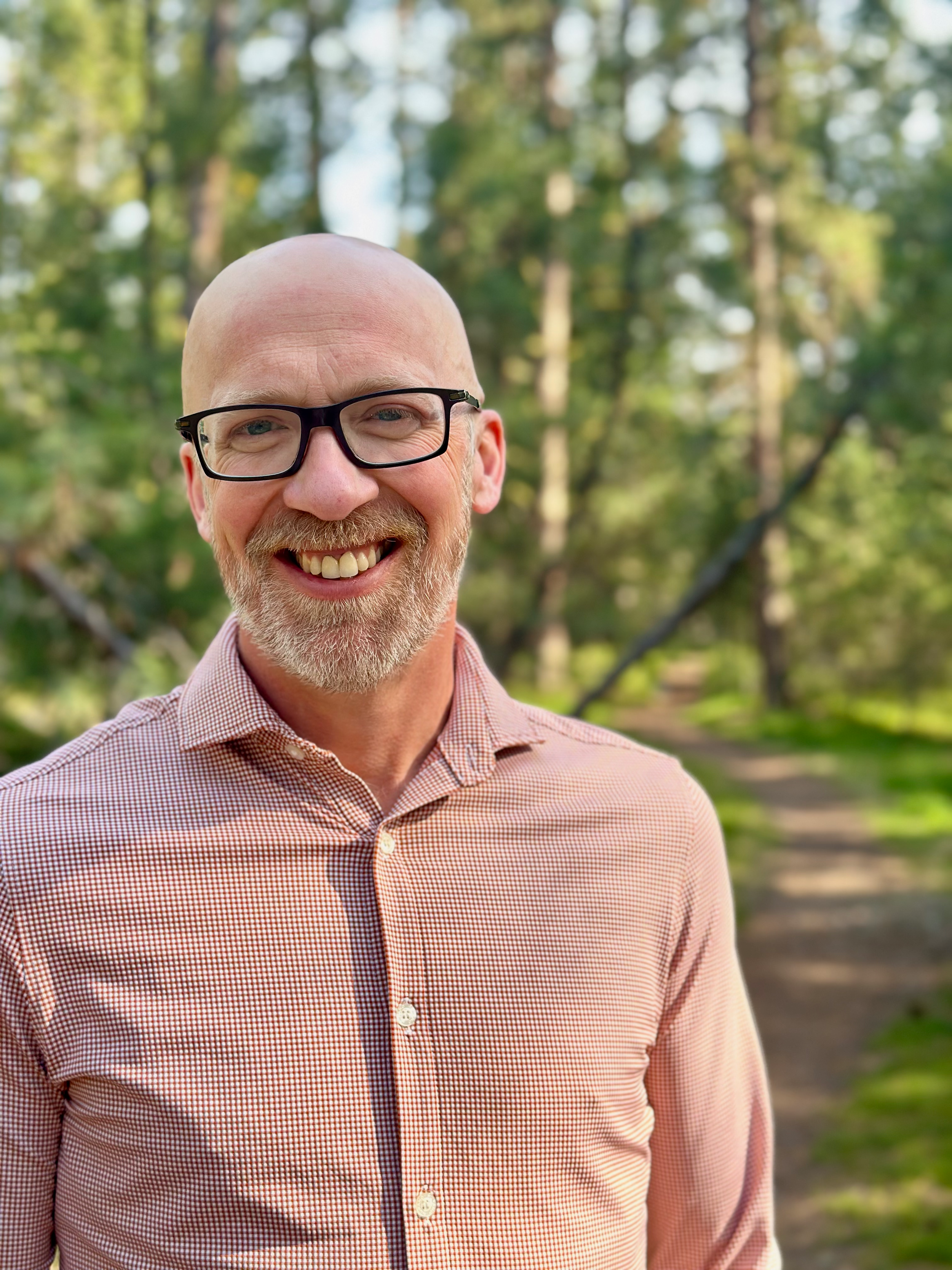 A smiling middle-aged man with a beard and glasses standing outdoors in a park with trees in the background.
