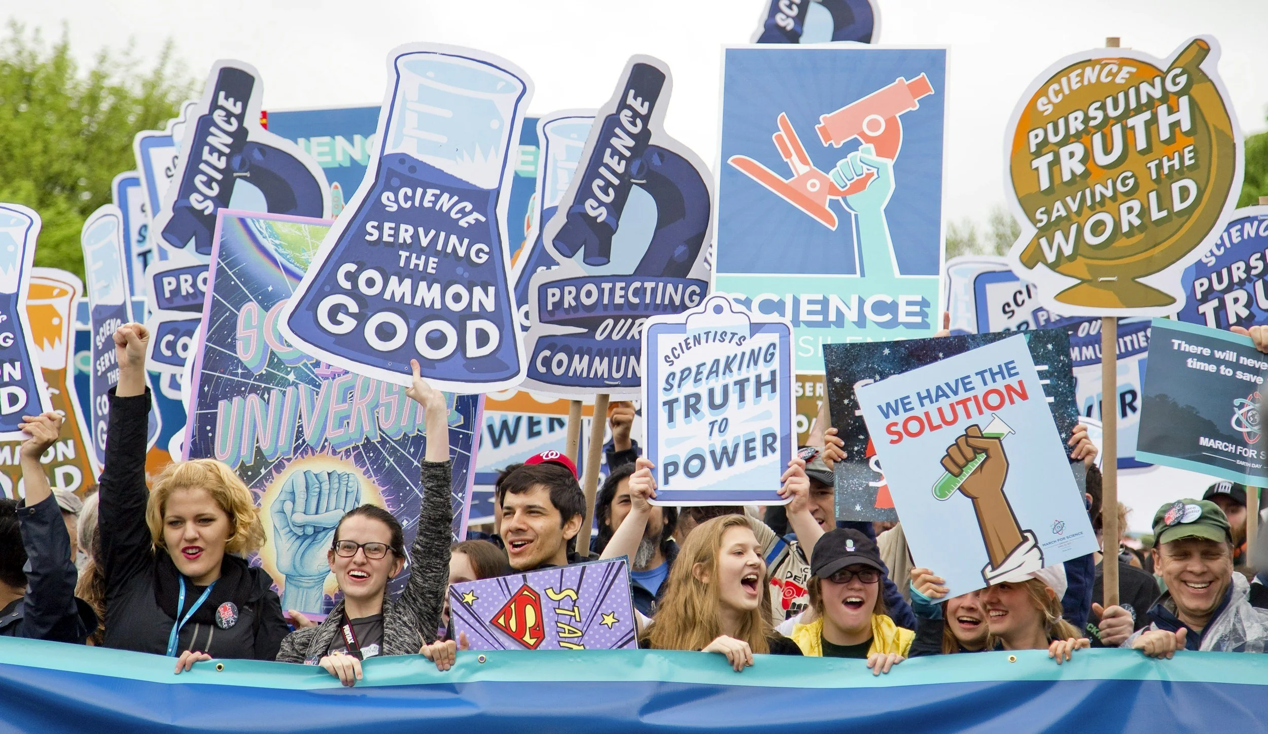 Group of people at a science march holding various signs promoting science, truth, and environmental protection, with some people smiling and cheering.