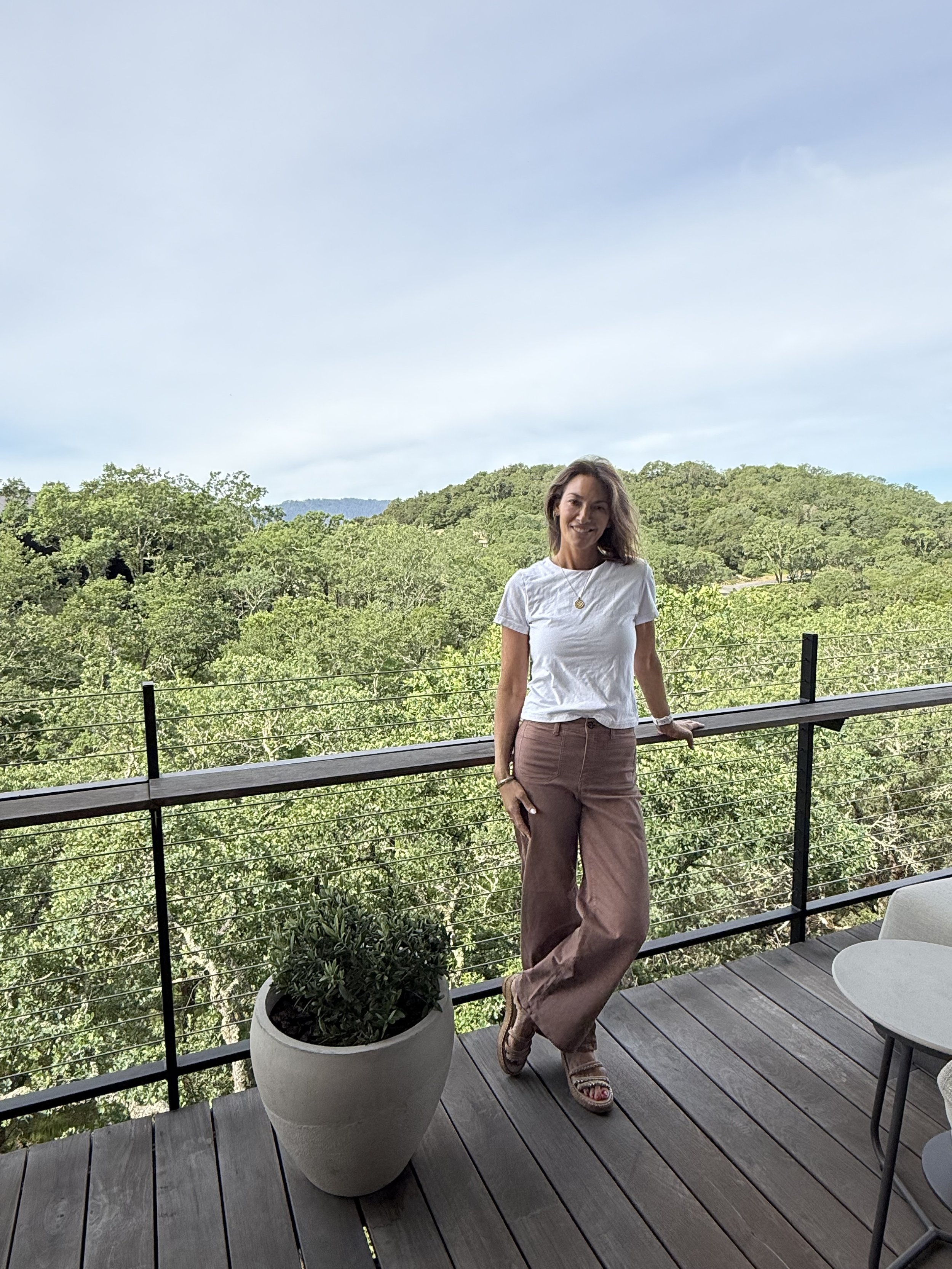 A woman standing on a wooden balcony with black railings, smiling and leaning lightly on the railing. She is wearing a white t-shirt, beige pants, and sandals. There is a large potted plant with green foliage in front of her. The background features a lush green forest and a partly cloudy sky.