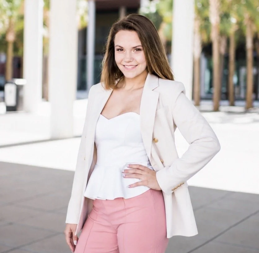 Young woman standing outdoors in professional attire, smiling with hand on hip, background of trees and modern building.