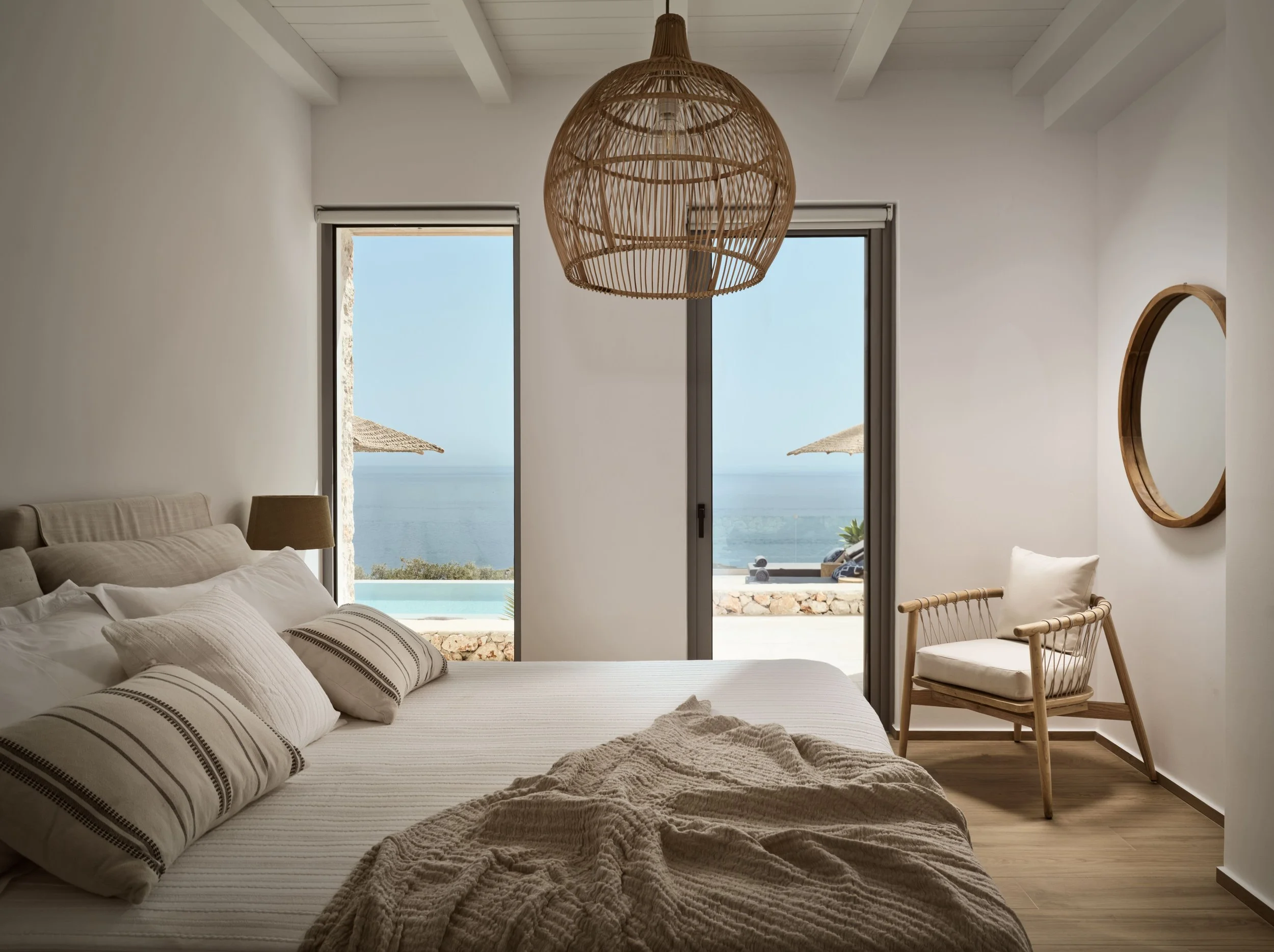 A bedroom with a large bed covered in white and beige striped bedding and pillows, a wooden chair, large glass doors with views of a balcony, ocean, and beach umbrellas outside, and a woven wicker pendant light hanging from the ceiling.