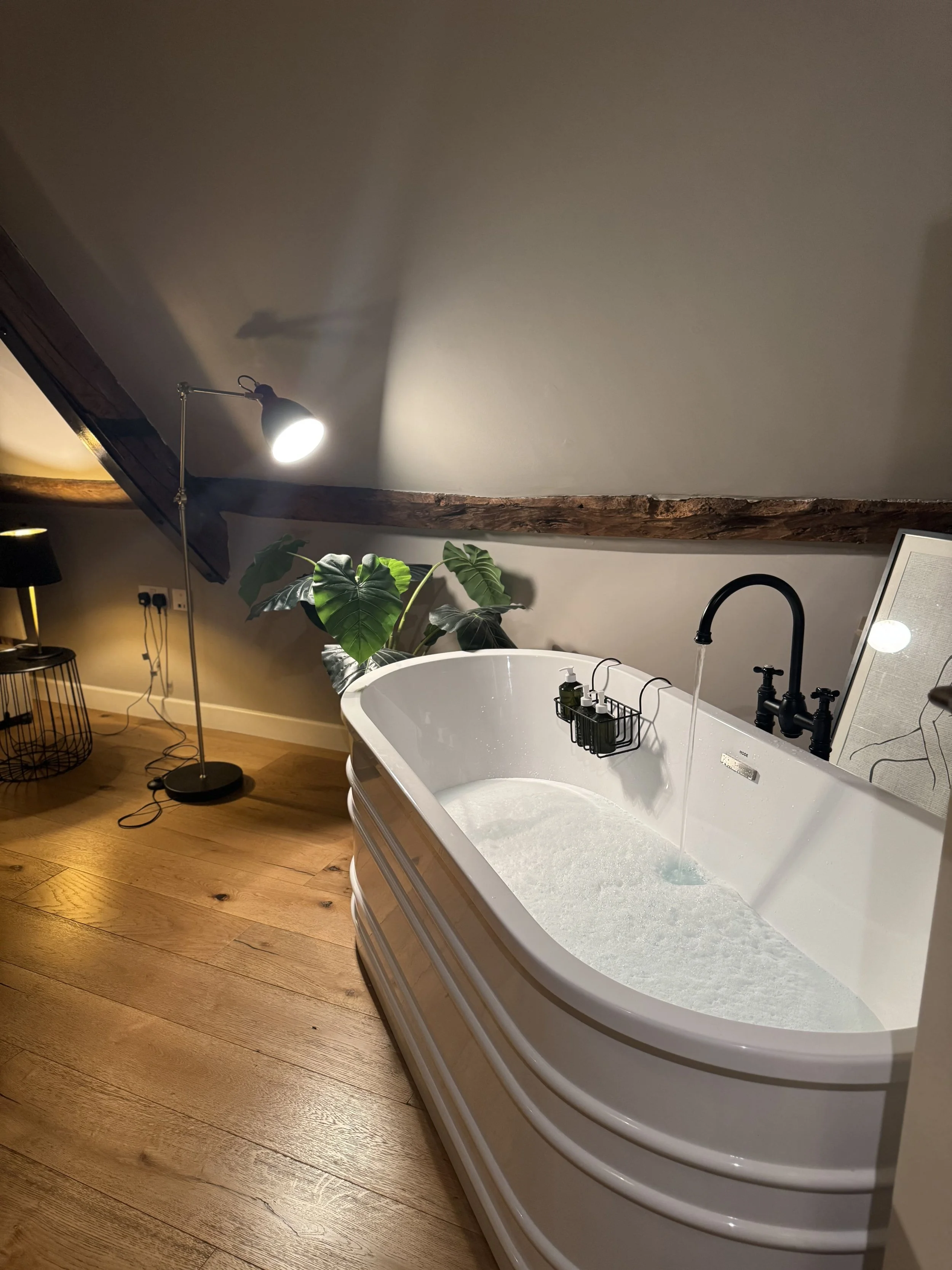 A bathroom with a white oval bathtub filled with water, a black faucet, a black showerhead, and a black metal rack holding soap and shampoo bottles. The bathroom has wooden floors, a large green plant, a black floor lamp, a framed picture on a stand,
