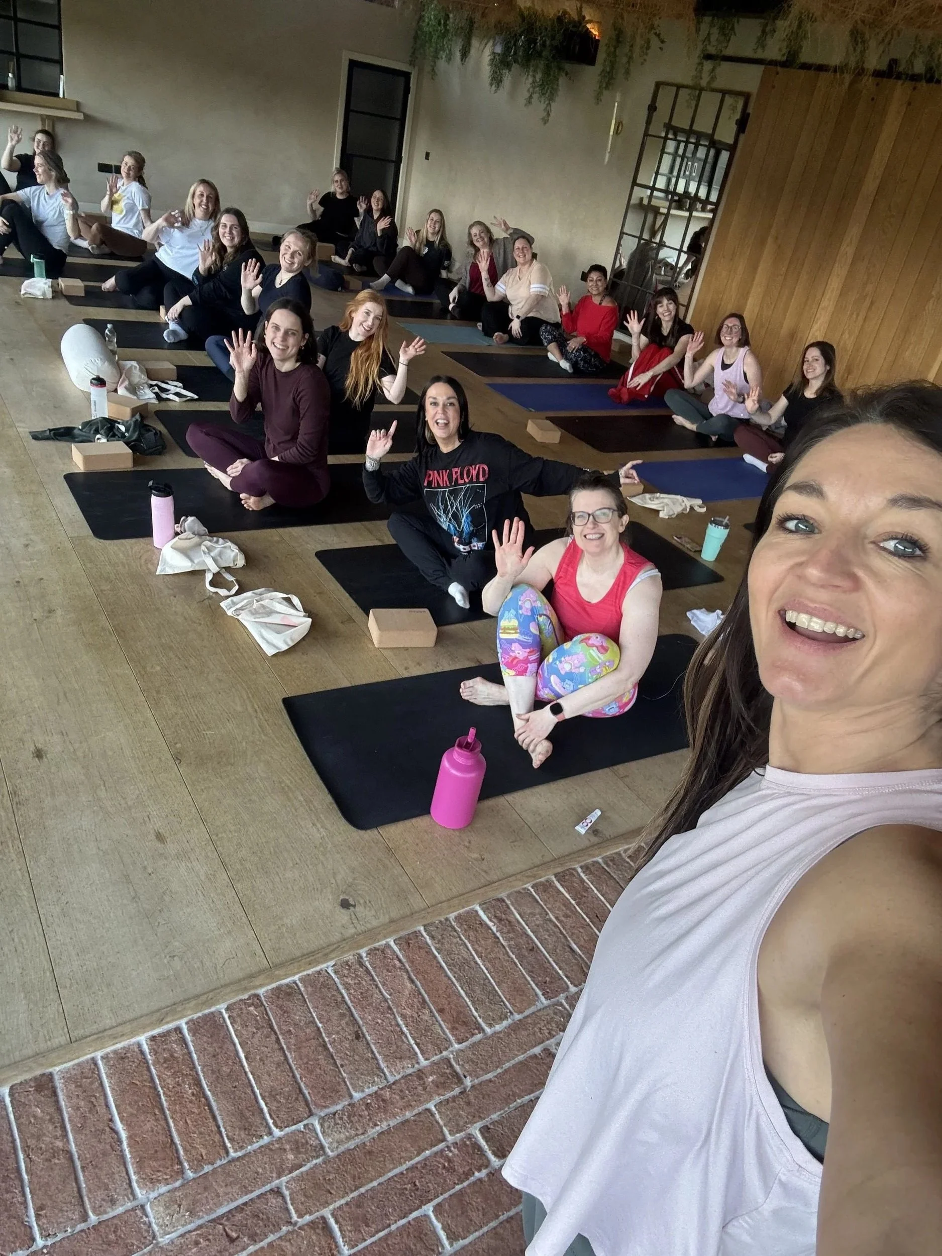 A group of women participating in a fitness class or yoga session in a studio with wooden walls. One woman in the foreground is taking a selfie, smiling, wearing a pink workout top and colorful leggings. The other women are sitting or stretching on m