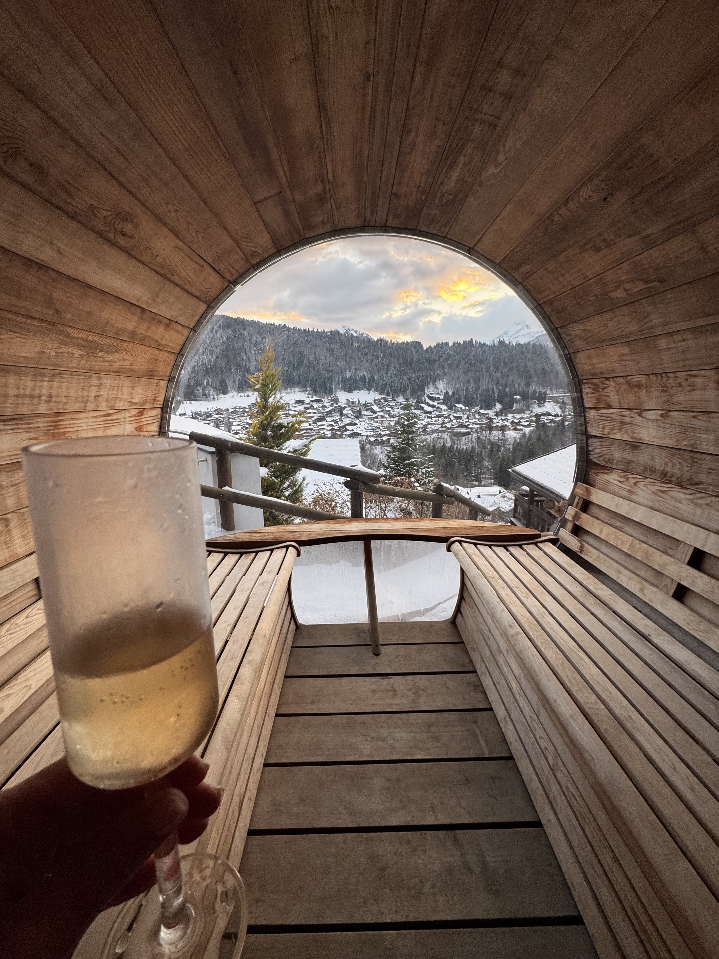 A person holding a glass of champagne or sparkling wine inside a wooden bubble-shaped sauna or relaxation pod overlooking a snowy mountain village and forest at sunset.