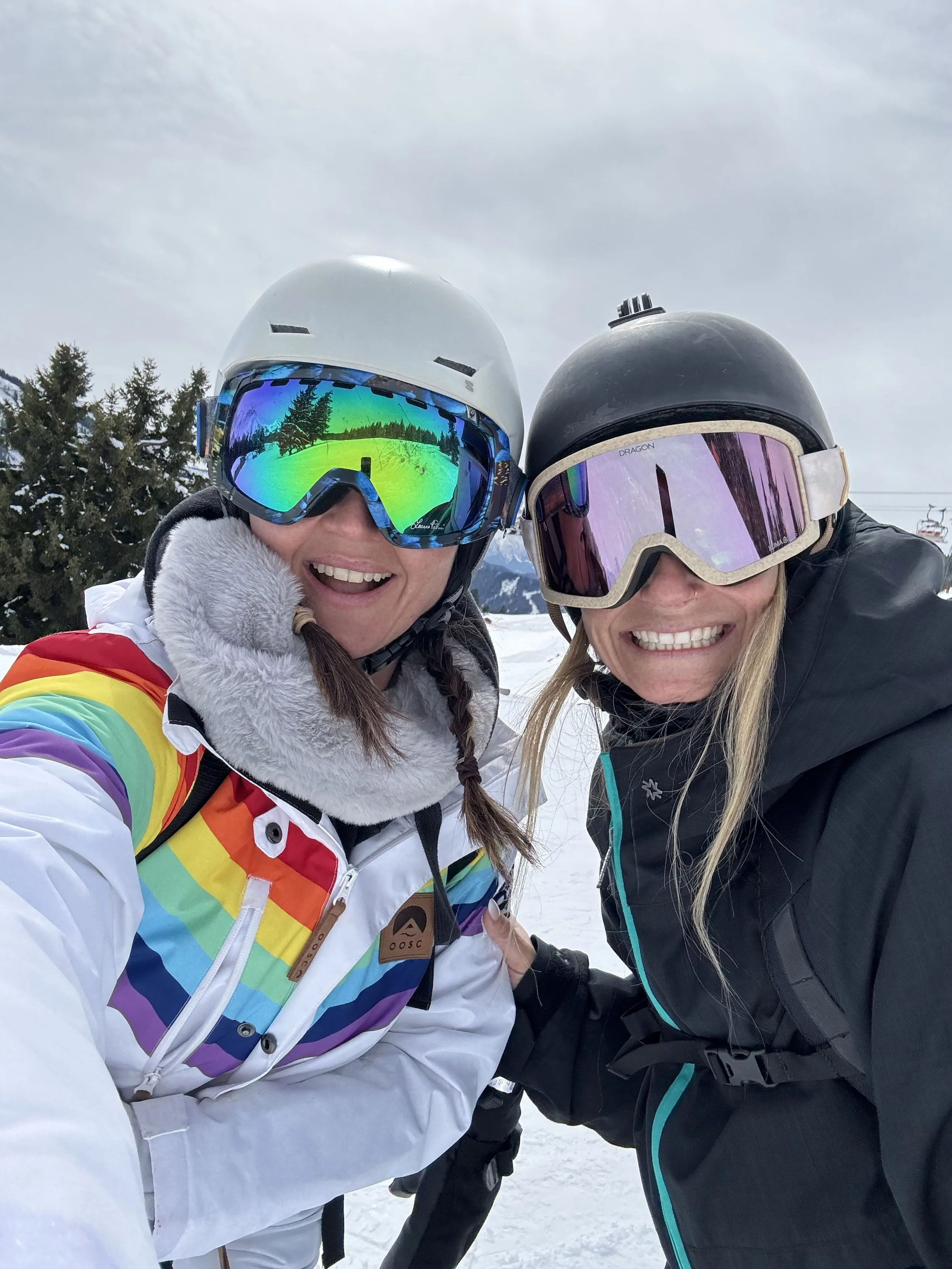 Two smiling women in ski gear and helmets on a snowy mountain, smiling at the camera with trees and ski lift in the background.