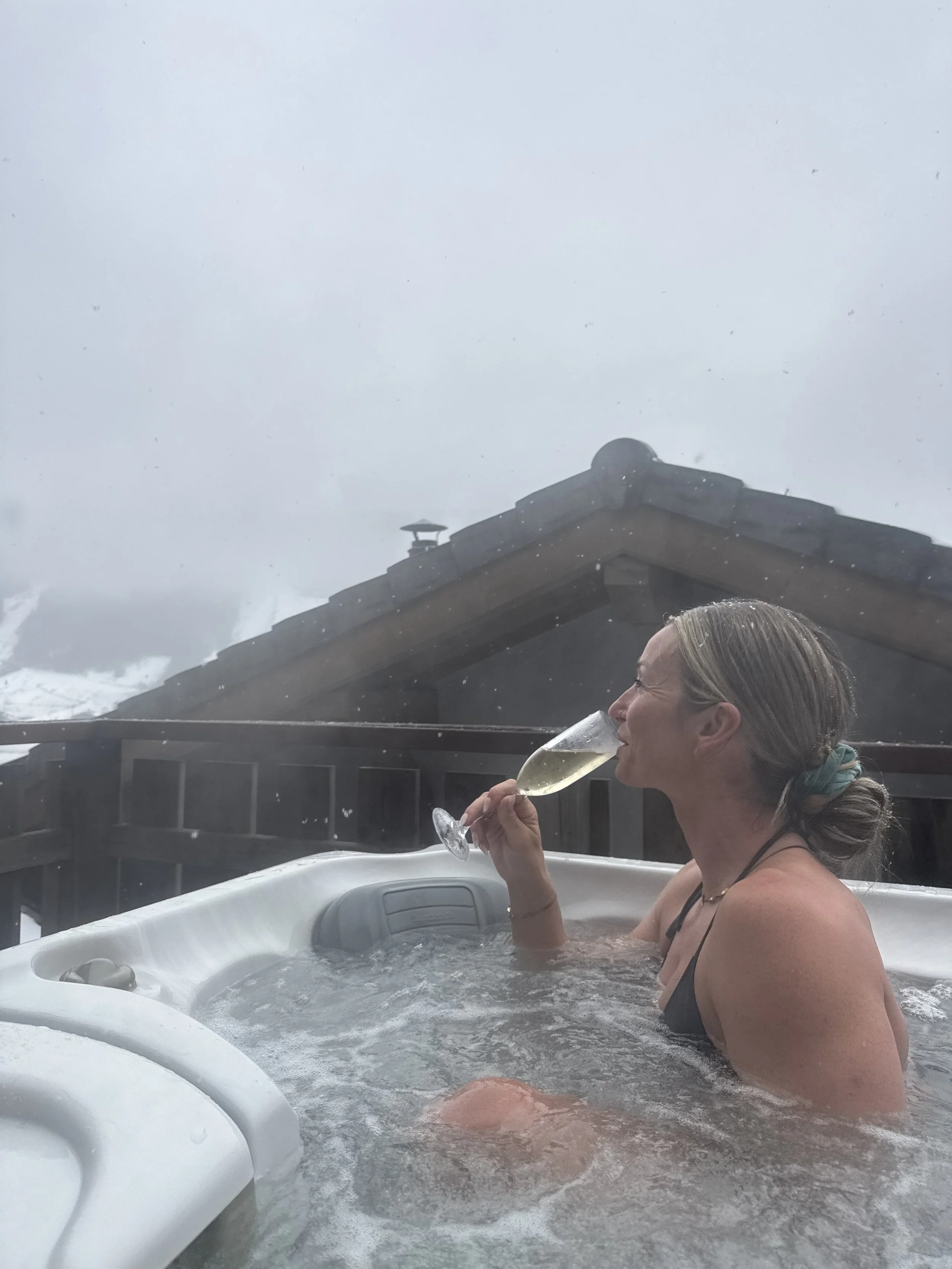 Woman relaxing in a hot tub on a snowy day, holding a glass of champagne, with snow falling and a house roof visible in the background.