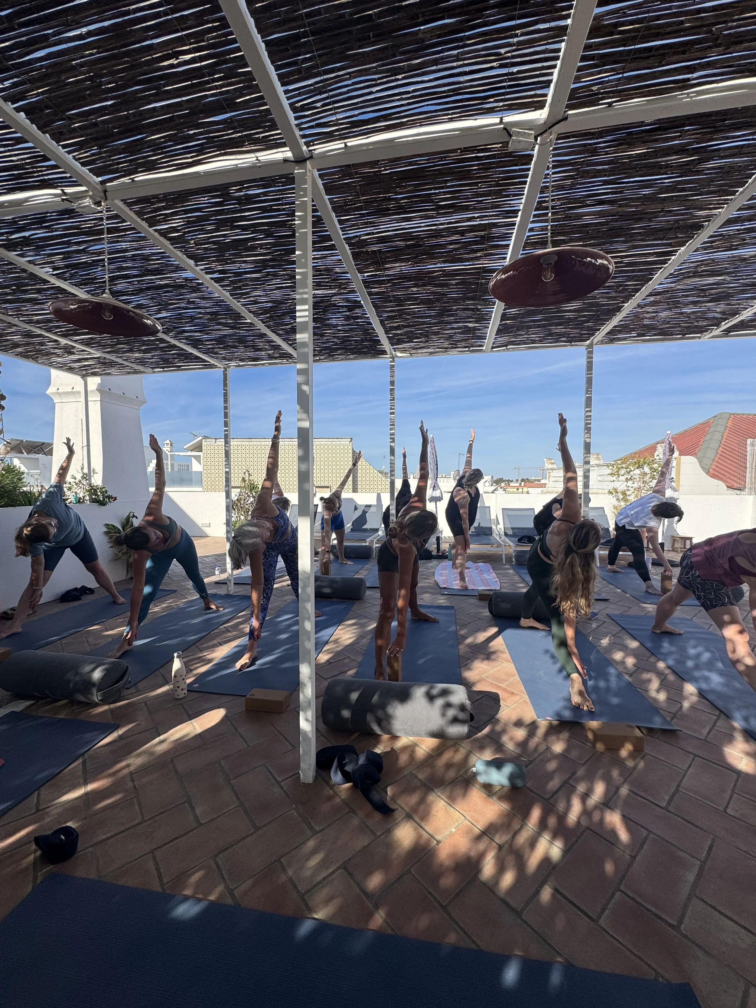 People practicing yoga outdoors under a pergola with a woven roof, on a terrace with potted plants, facing blue sky and white buildings in the background.
