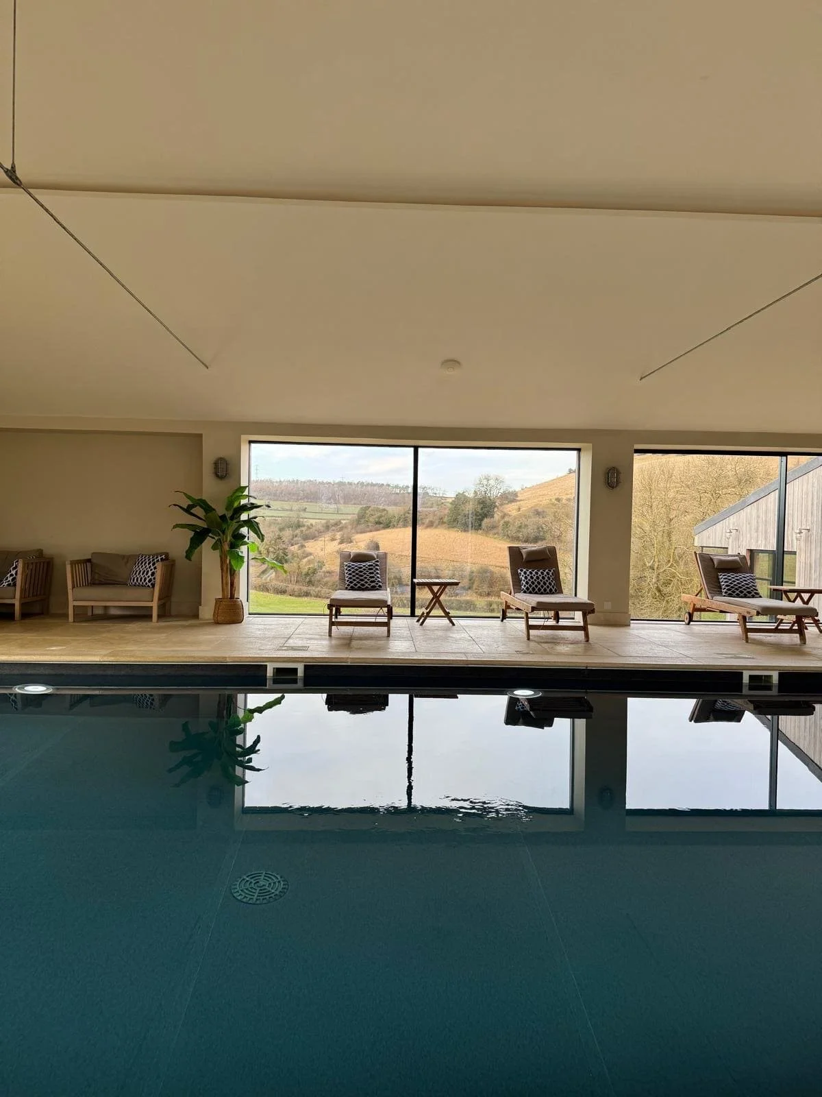 Indoor swimming pool area with lounge chairs and large window showing a scenic countryside view.