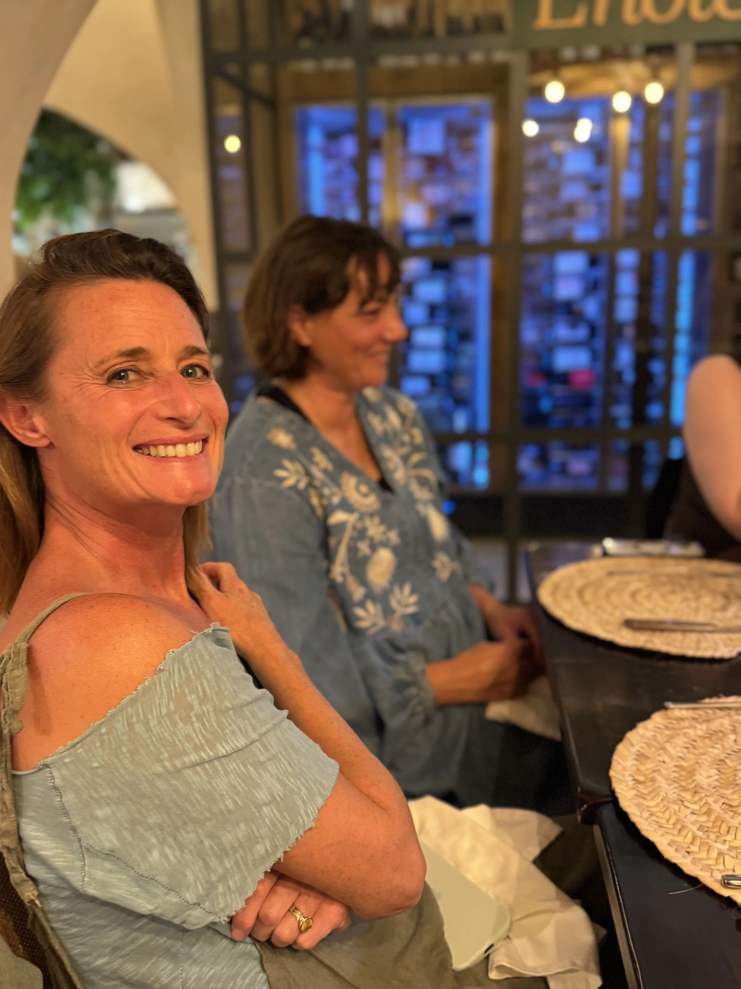 Smiling woman with shoulder-length brown hair, wearing a light gray off-shoulder top, sitting at a table with woven placemats. In the background, another woman with short brown hair, blue patterned blouse, and a blurred restaurant setting with warm l