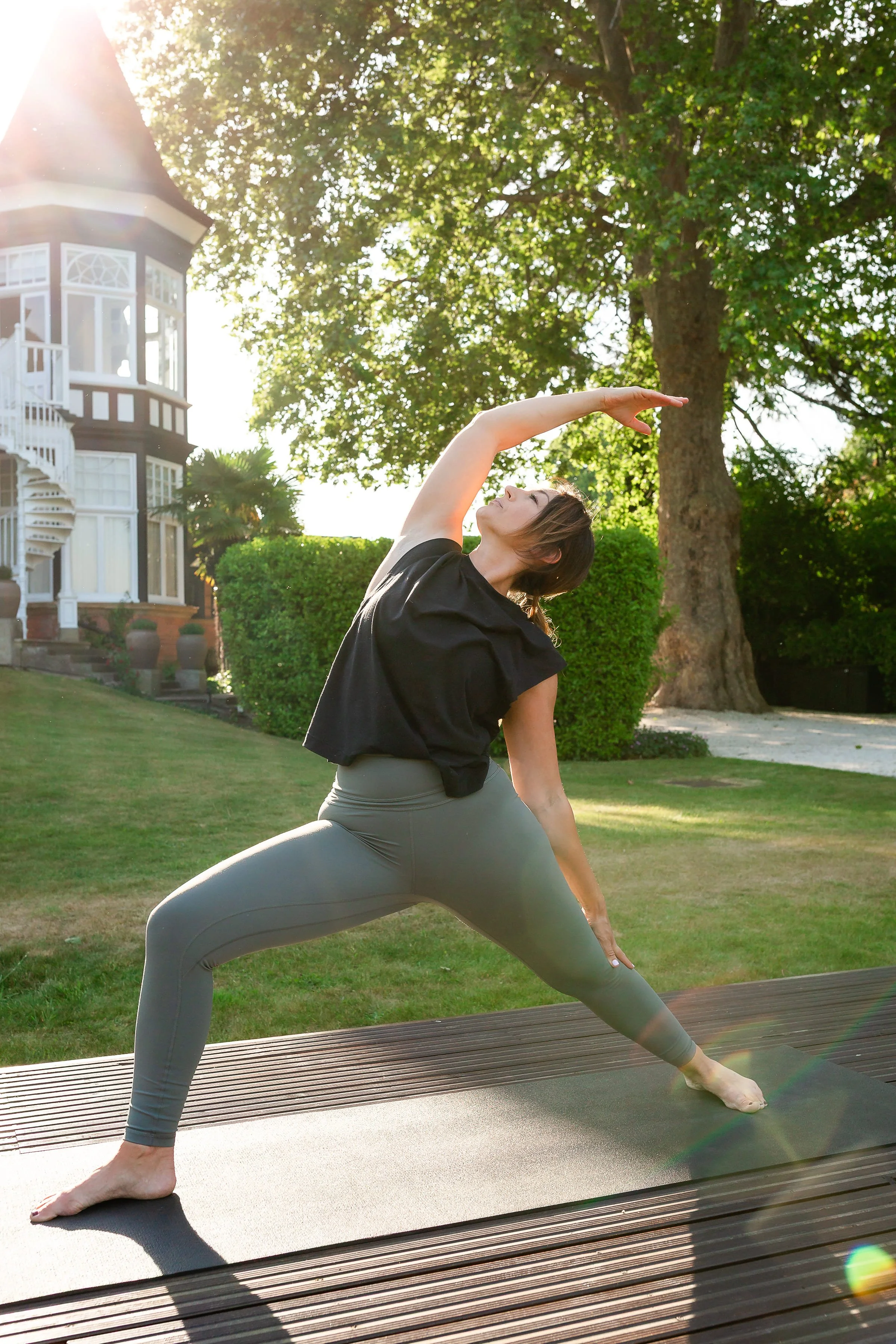 A woman practicing yoga outdoors on a mat in a garden, wearing a black top and gray leggings, in a wide lunge pose with one arm extended overhead and the other resting on her thigh, with trees and a house in the background.