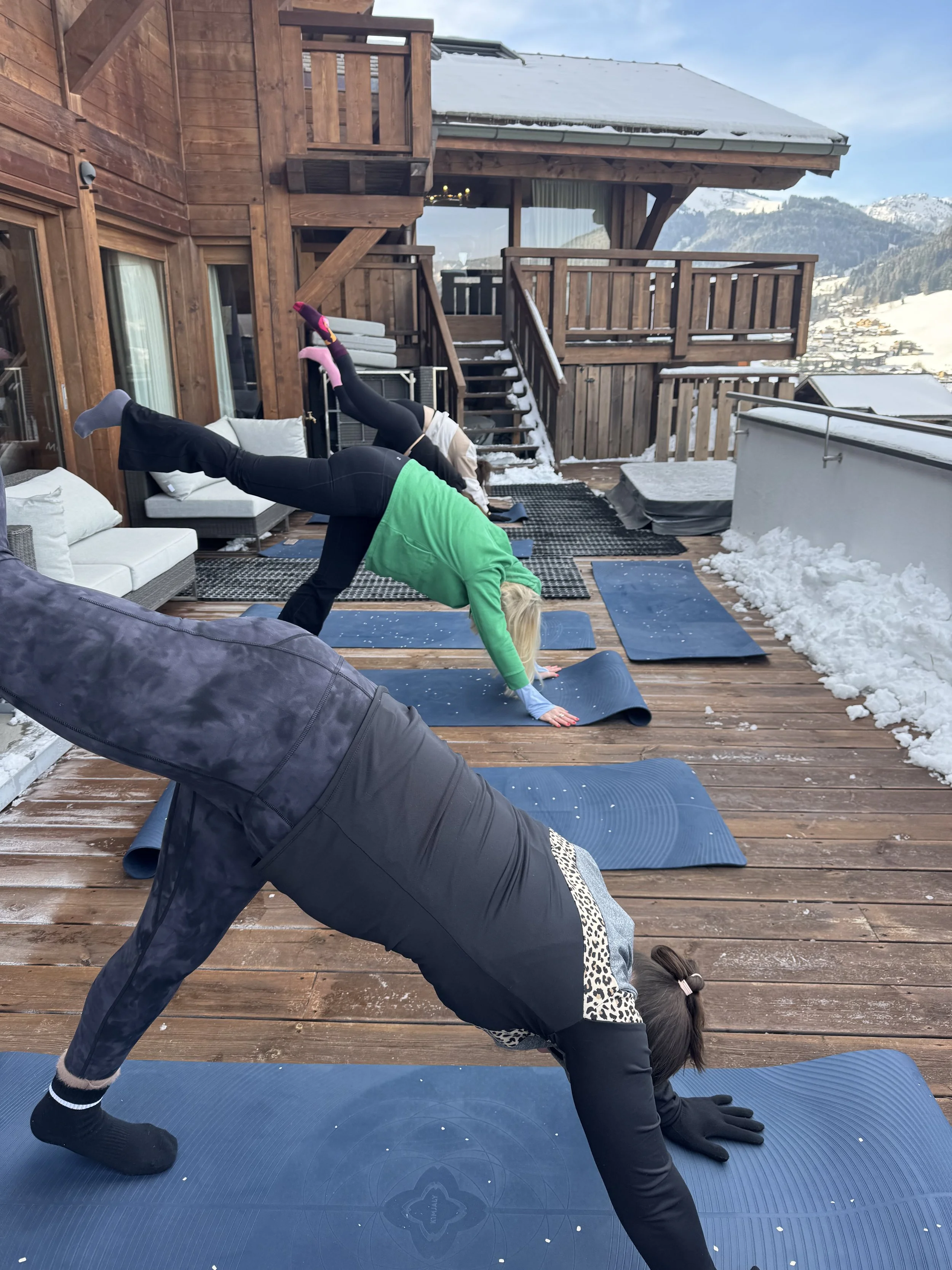 People practicing yoga outdoors on a wooden deck with snow, mountains, and sky in the background.
