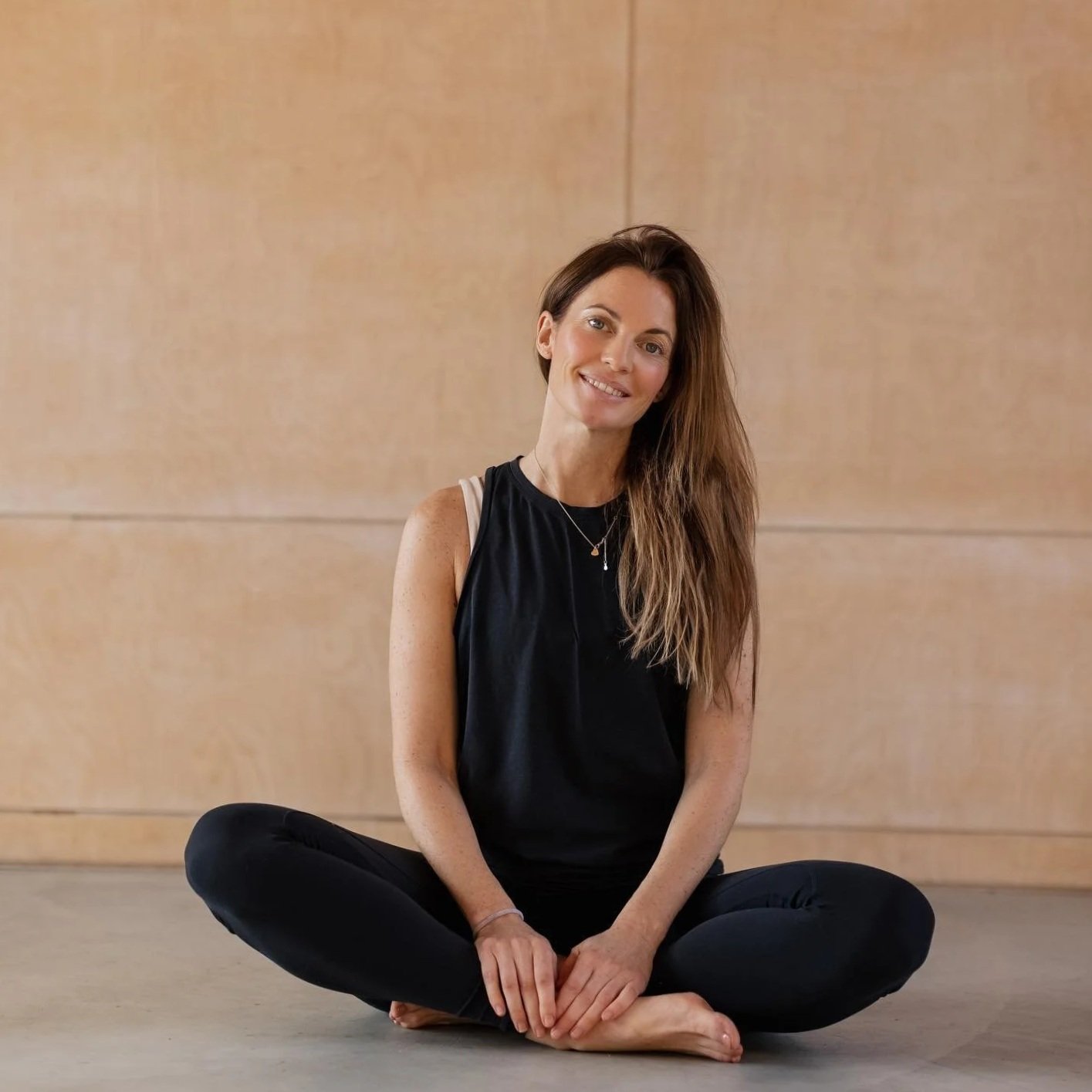 A woman with long brown hair sitting cross-legged on the floor in front of a beige wall, smiling at the camera, wearing a black tank top and black pants.