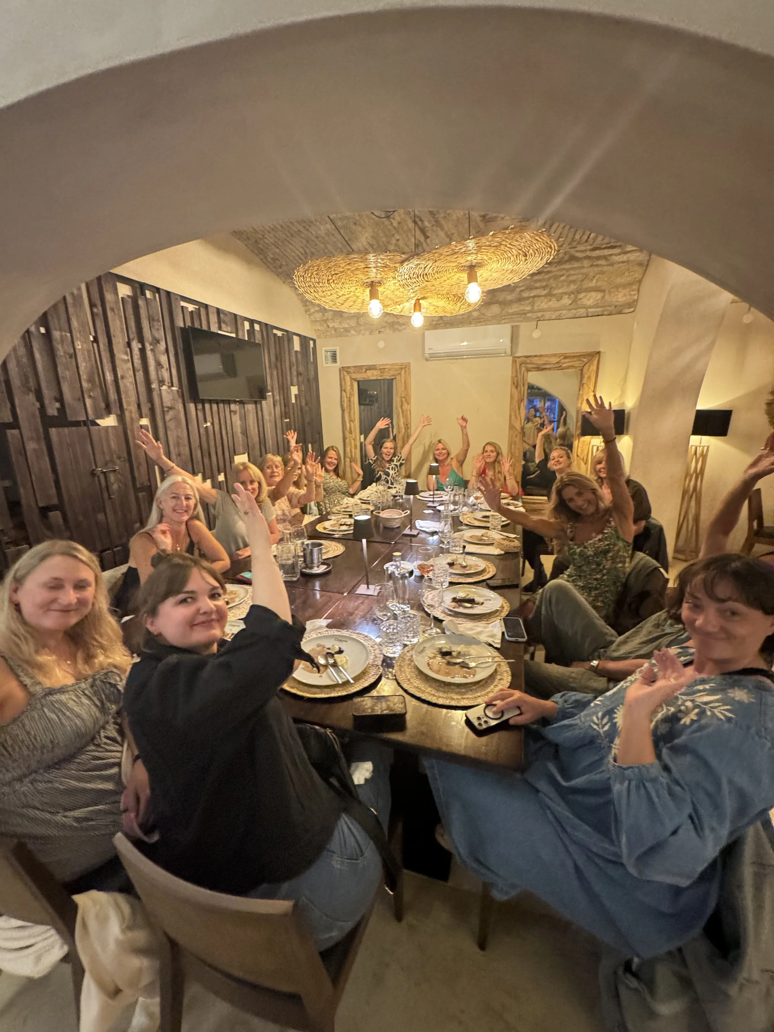 A group of people seated around a large dining table, smiling and raising their hands in celebration, in a warmly lit room with modern decor.
