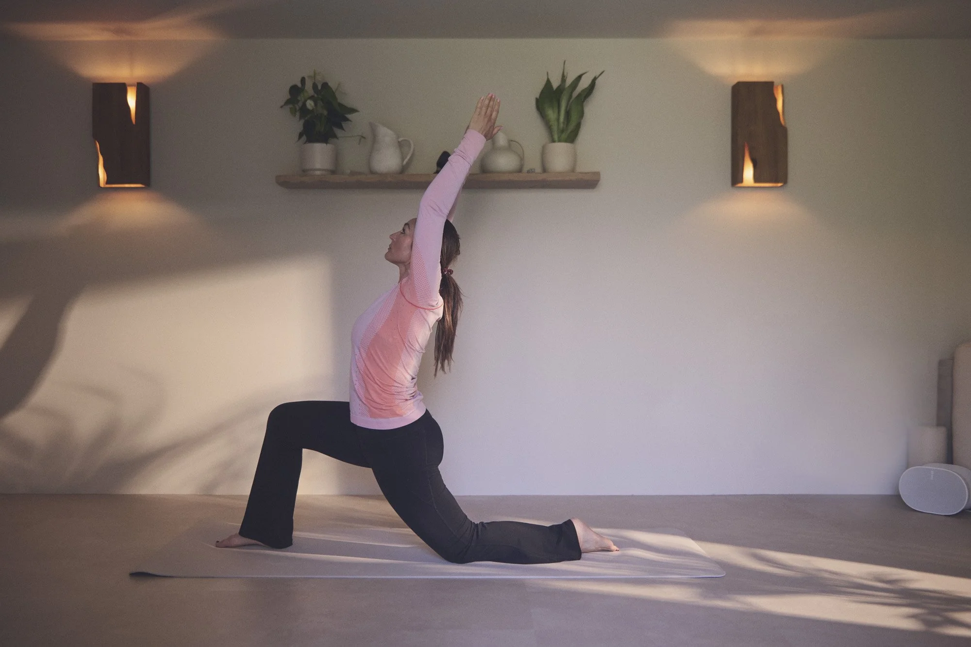 A woman practicing yoga indoors, performing a low lunge pose on a mat, with her hands raised above her head. The room is softly lit with wall-mounted lights and decorated with potted plants on a shelf.