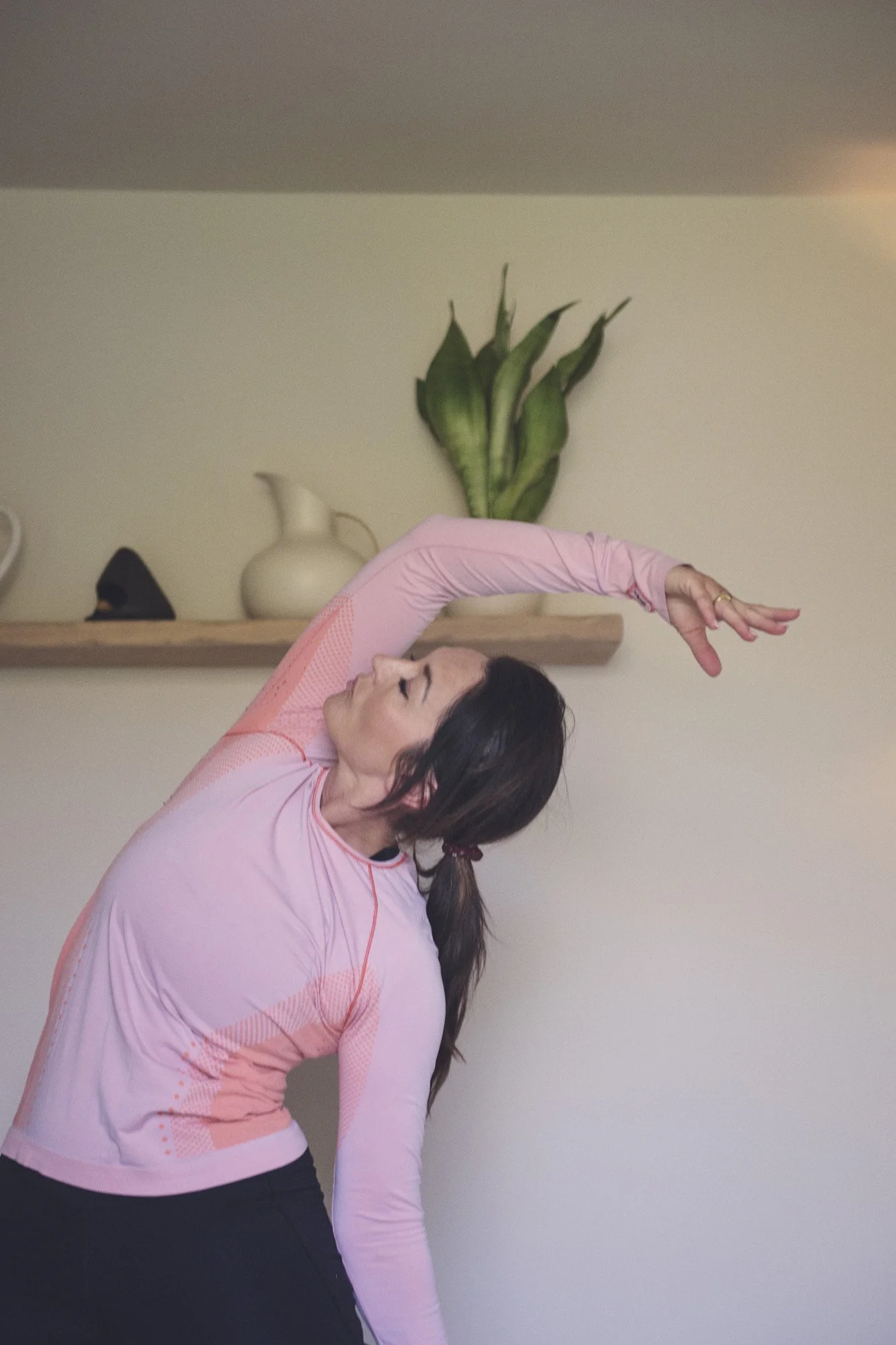 A woman with dark hair extended backwards, wearing a light pink, long-sleeve athletic shirt, bending her head and torso to the side, with an arm raised above her head.