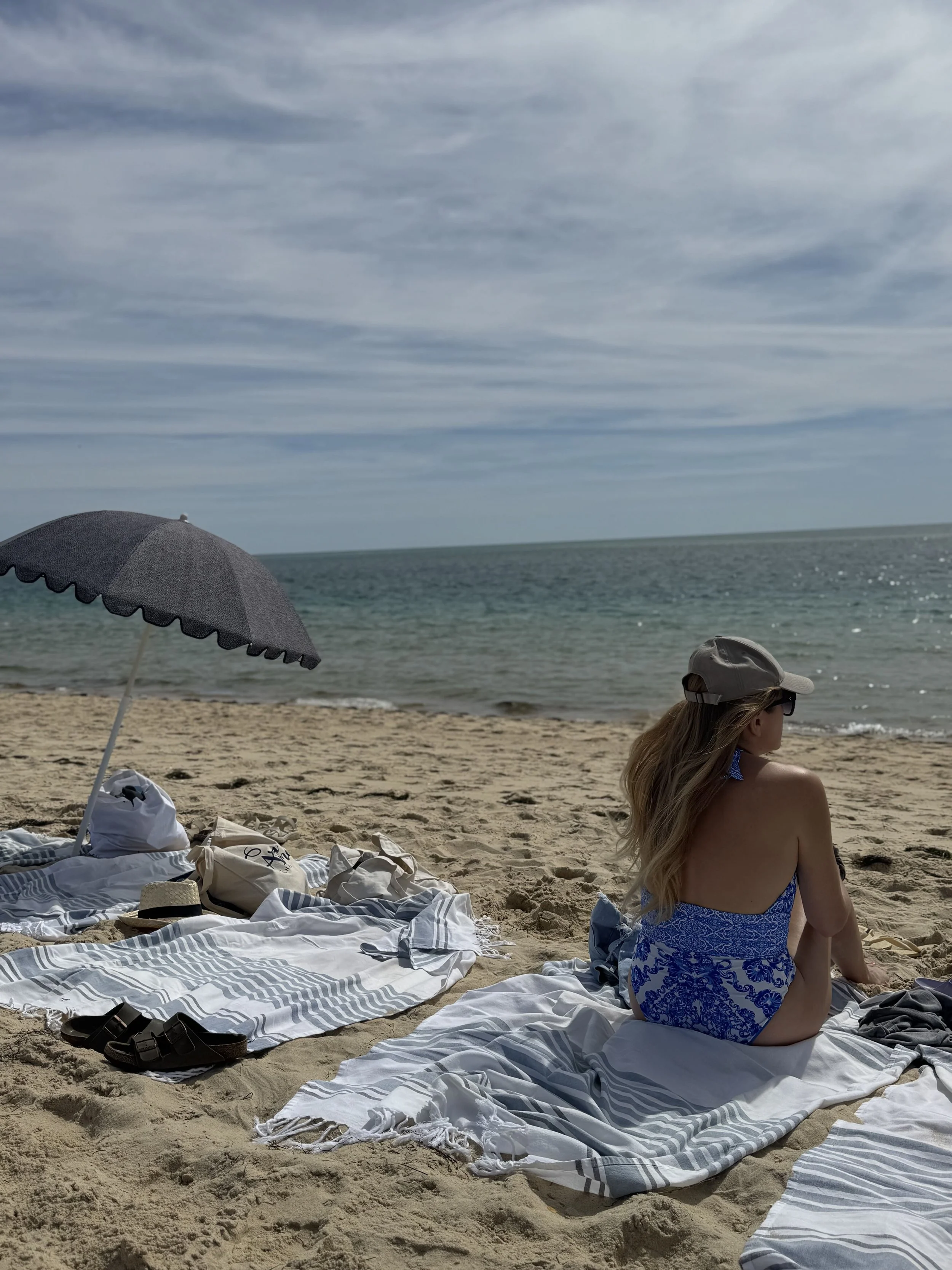 A woman sitting on a striped towel at the beach, facing the water. There is a large gray umbrella and some bags and shoes nearby, with a cloudy sky and ocean in the background.