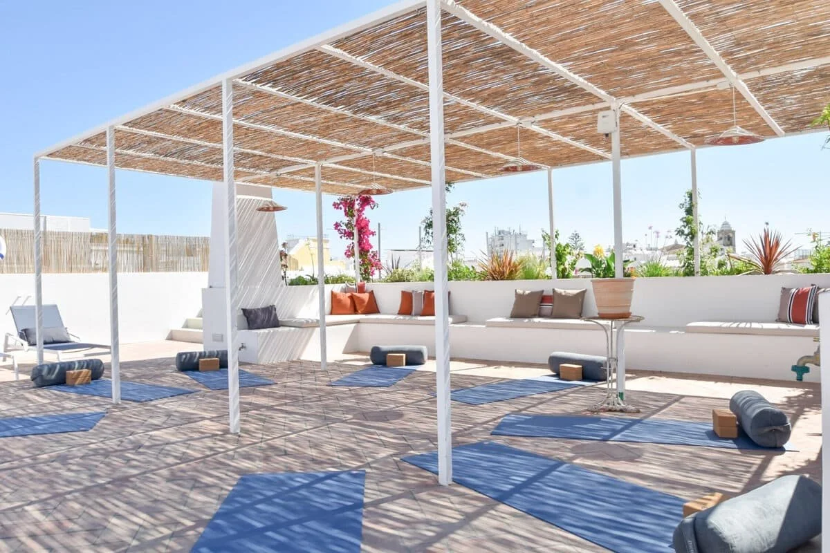 Outdoor yoga or relaxation space with blue mats, pillows, and foam rollers, shaded by a bamboo ceiling, with plants and white seating along the back wall.