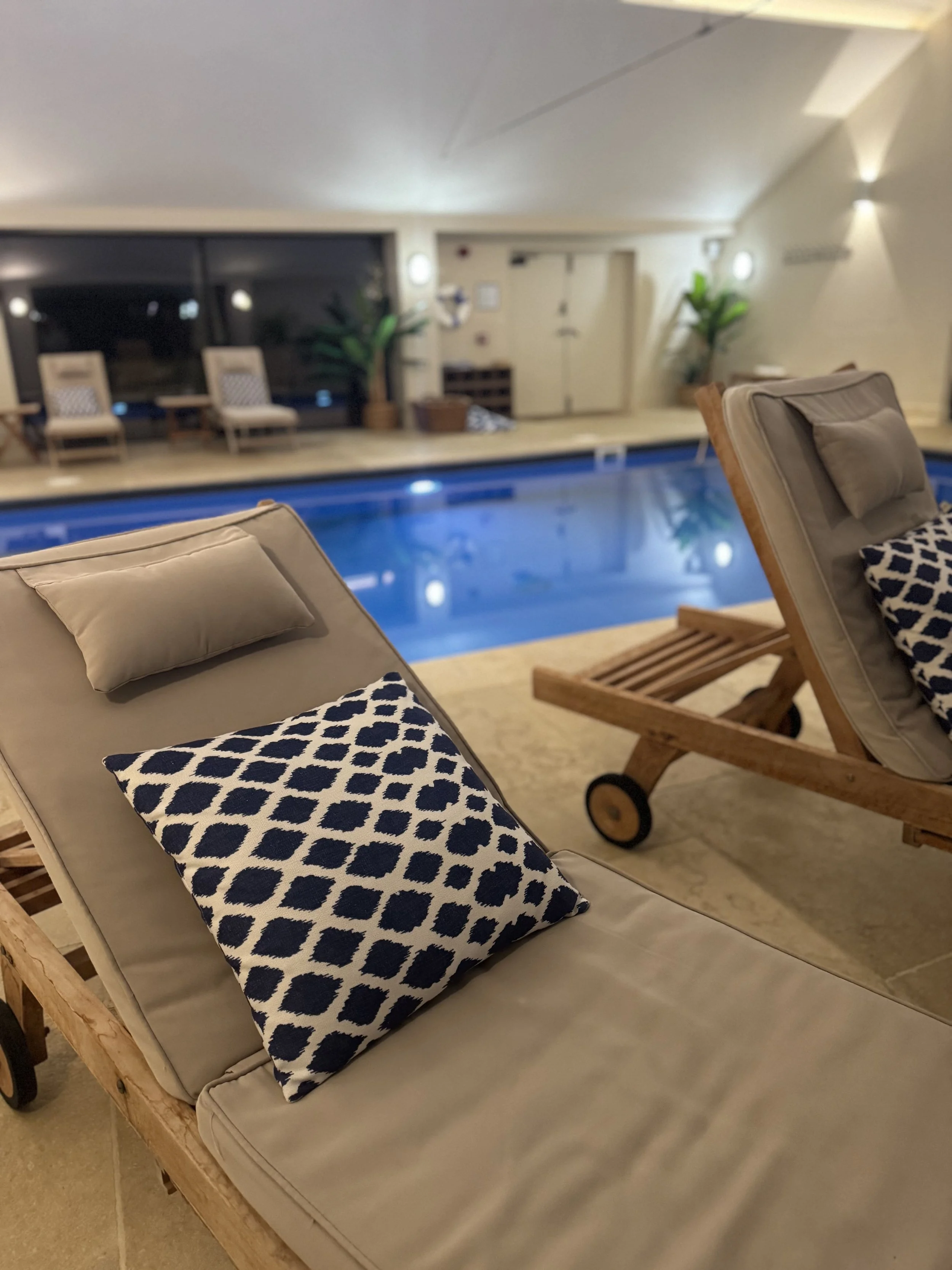 Indoor pool area with beige lounge chair with black and white patterned pillow, wooden lounge chair, and potted plants, with seating area and a large sliding glass door in the background.