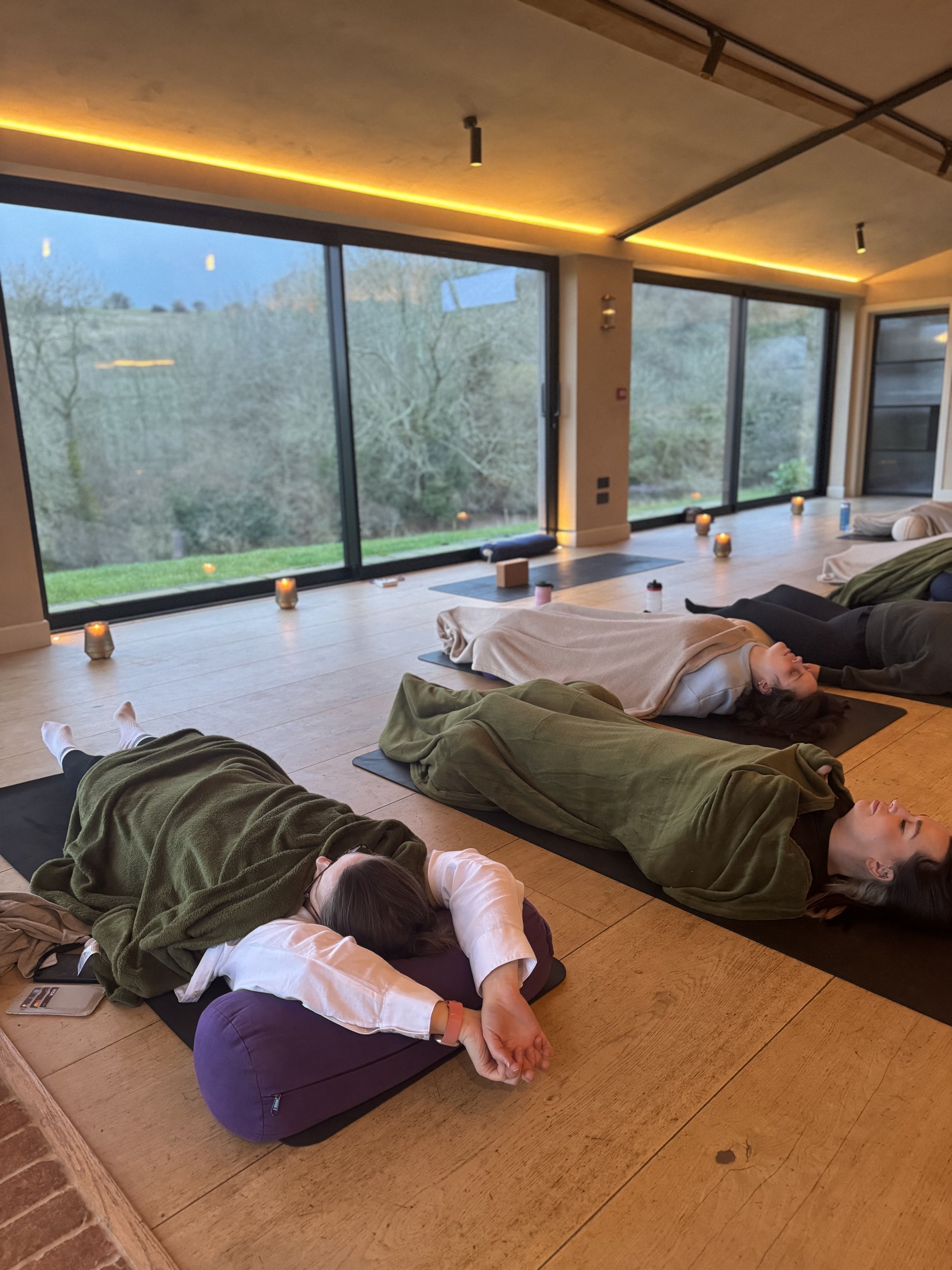 People lying on yoga mats in a peaceful indoor setting with large windows, candles, and serene outdoor view.