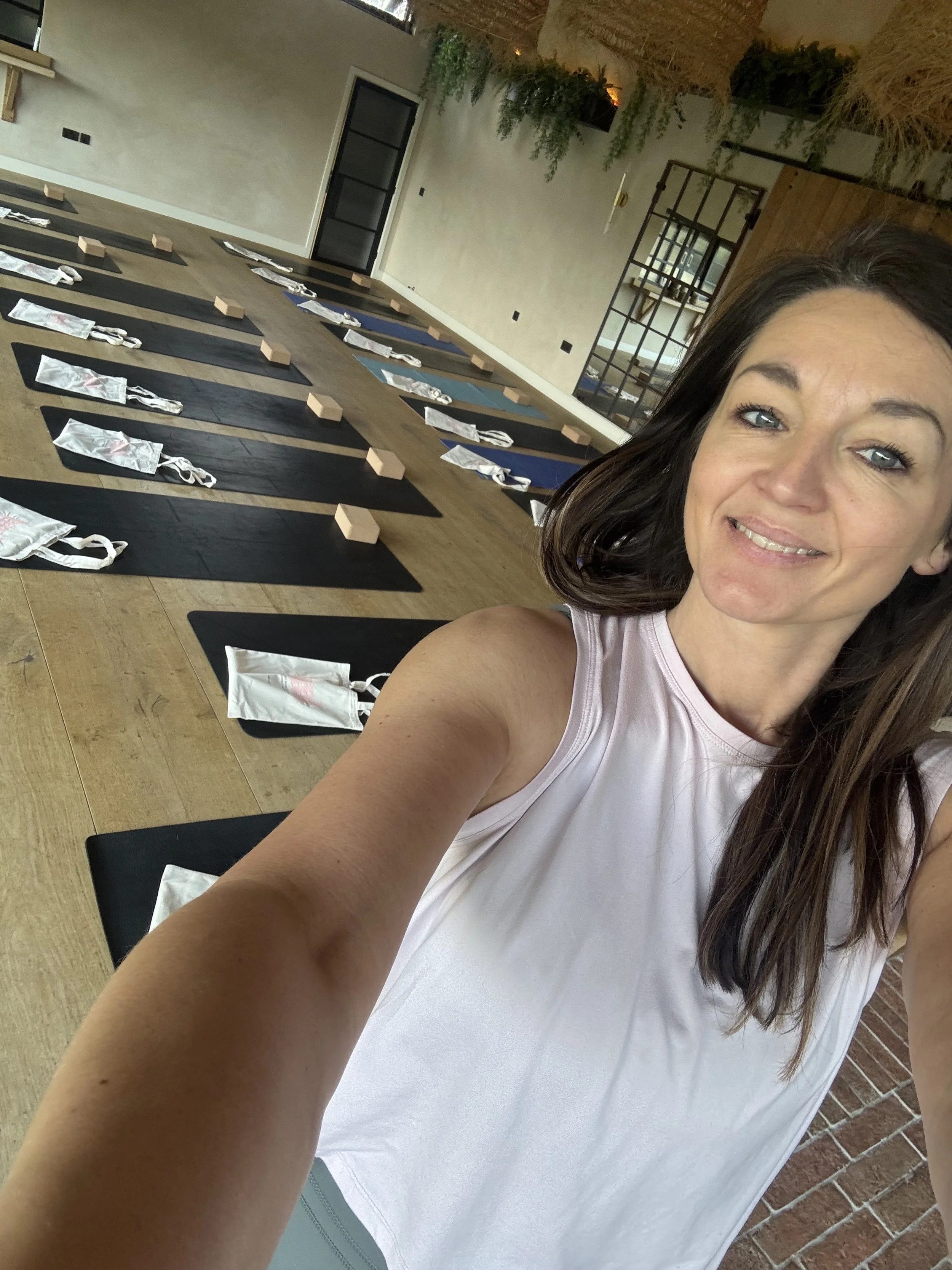 A woman taking a selfie in a fitness or yoga studio with black mats, water bottles wrapped in plastic, and small wooden blocks on the mats. The studio has wooden flooring, a brick wall, and a door with glass panes.