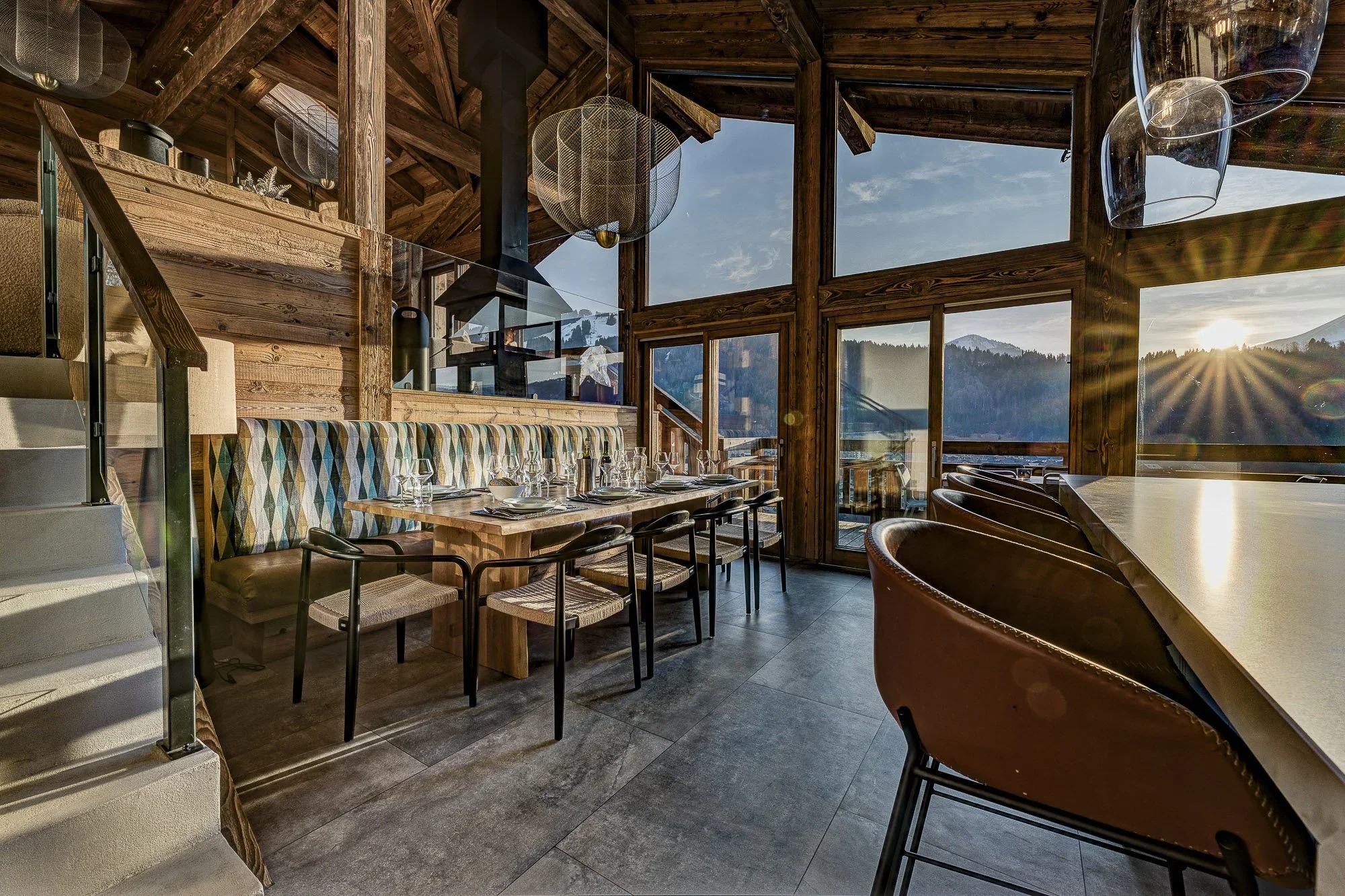 Interior of a rustic restaurant with wooden walls and large windows showing mountain views, decorated with modern wire, glass pendant lights, set tables with glassware, and leather chairs.