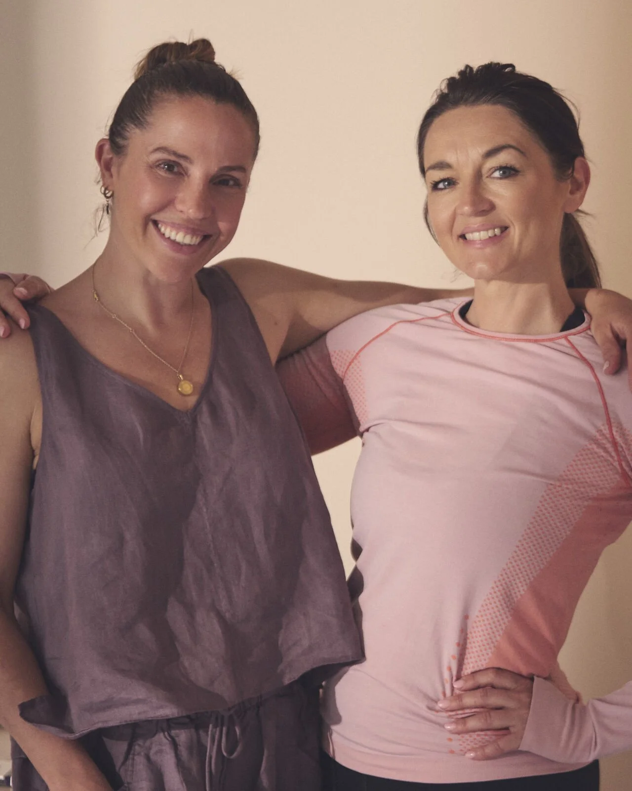 Two women smiling with their arms around each other, standing against a plain background.