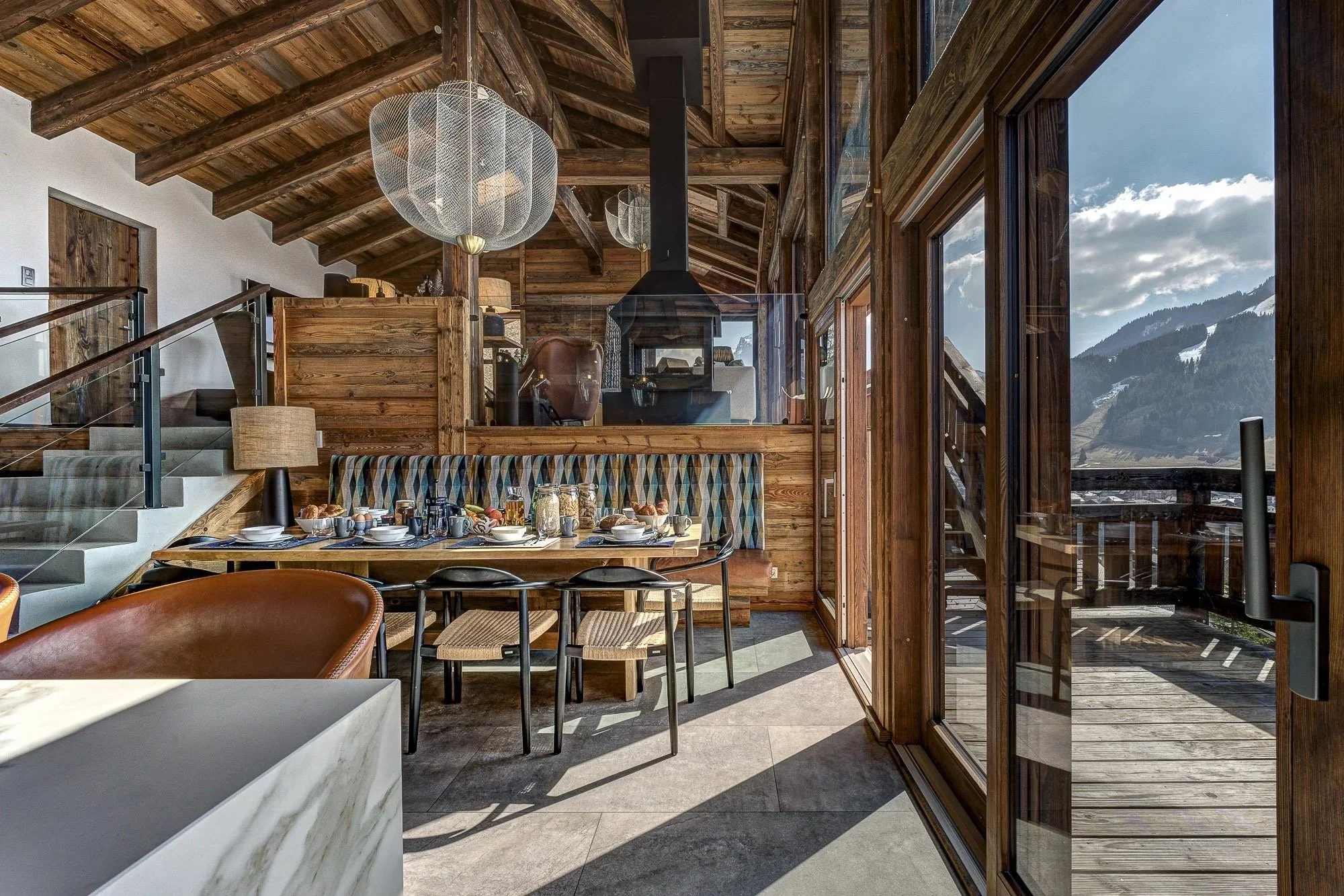 Interior of a modern house with wood accents, large windows, and mountain views, featuring a dining table set for breakfast.