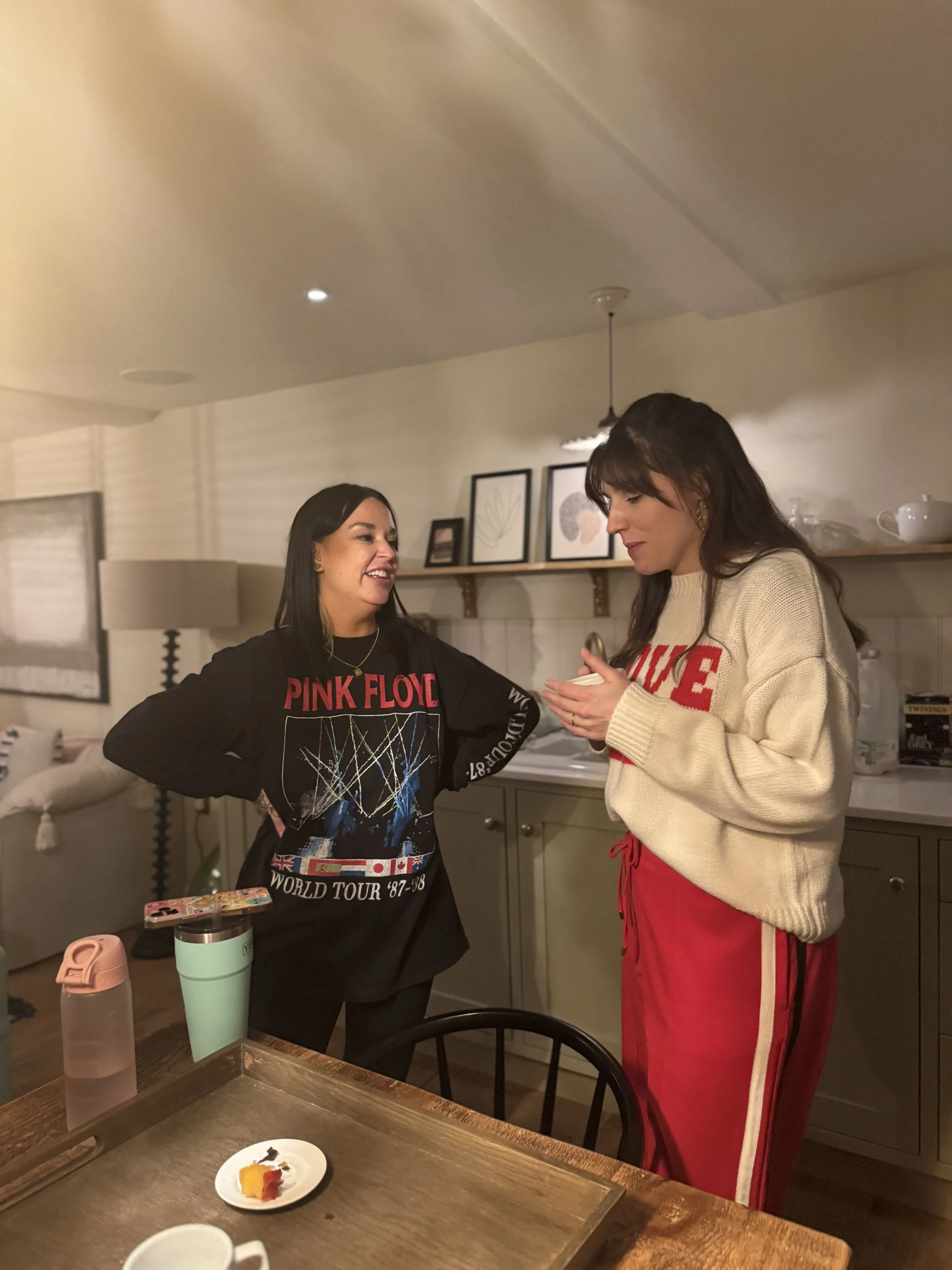 Two women standing and engaging in conversation in a kitchen, one wearing a Pink Floyd sweatshirt, and the other in a beige sweater and red pants, with a sailboat model and decorative framed art on a shelf behind them.