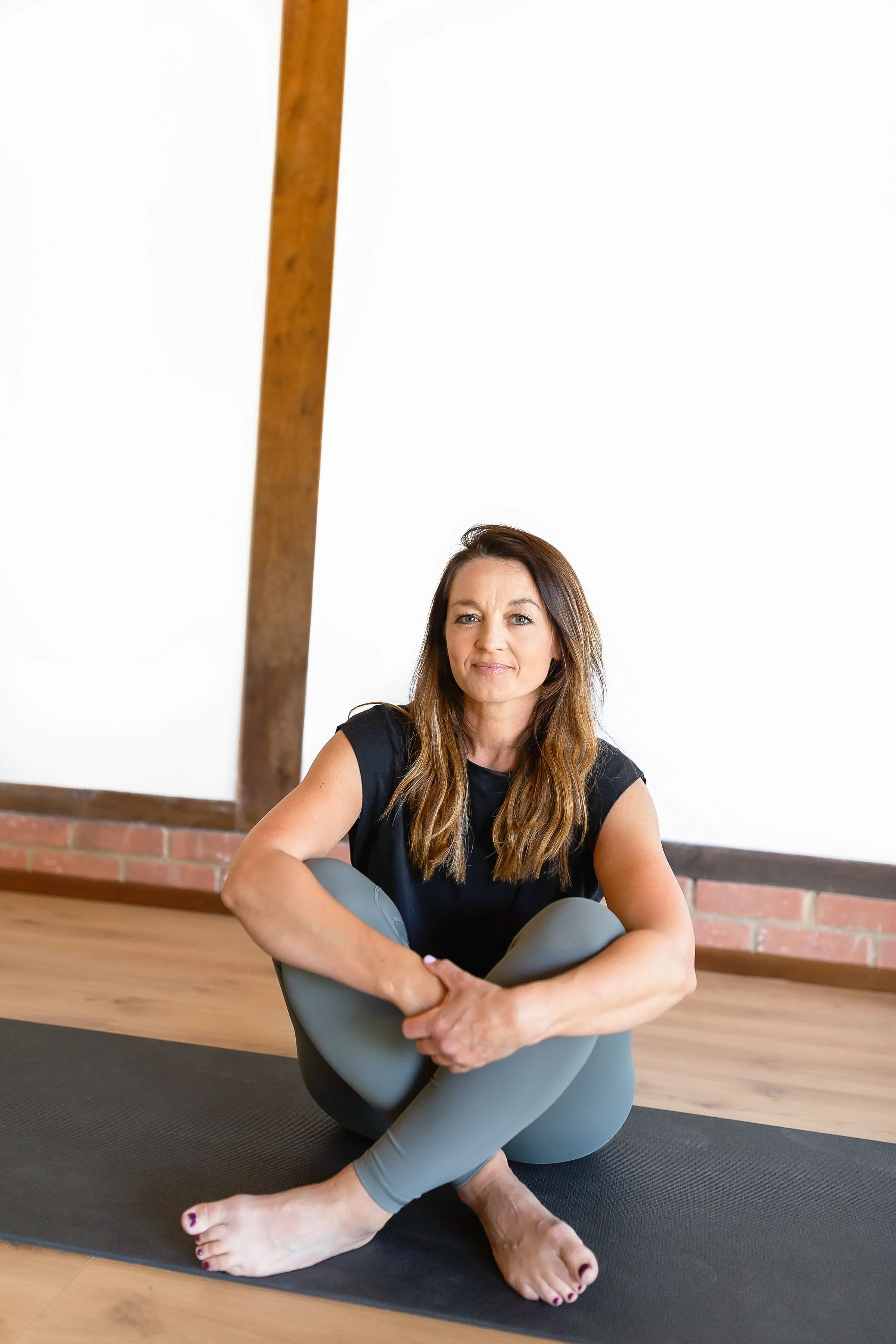 A woman sits on a yoga mat in a yoga studio, holding her knees while crossing her arms around her legs. She has long brown hair, wears a black t-shirt and gray leggings, and is in a relaxed, calm pose.