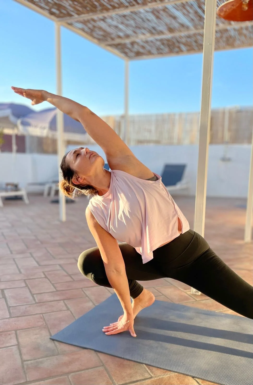 A woman practicing yoga outdoors on a mat, performing a side stretch with her left arm extended overhead, during sunlight with a patio and lounge chairs in the background.