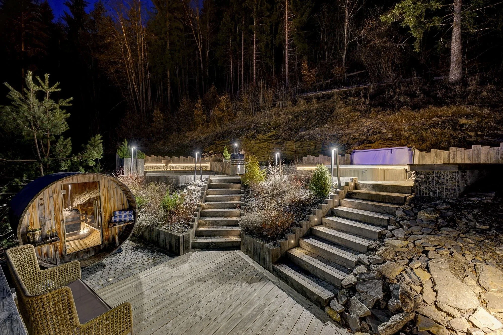 Nighttime view of a backyard patio with wooden stairs, a small covered hot tub, outdoor chairs, and a forested hillside in the background illuminated by outdoor lights.