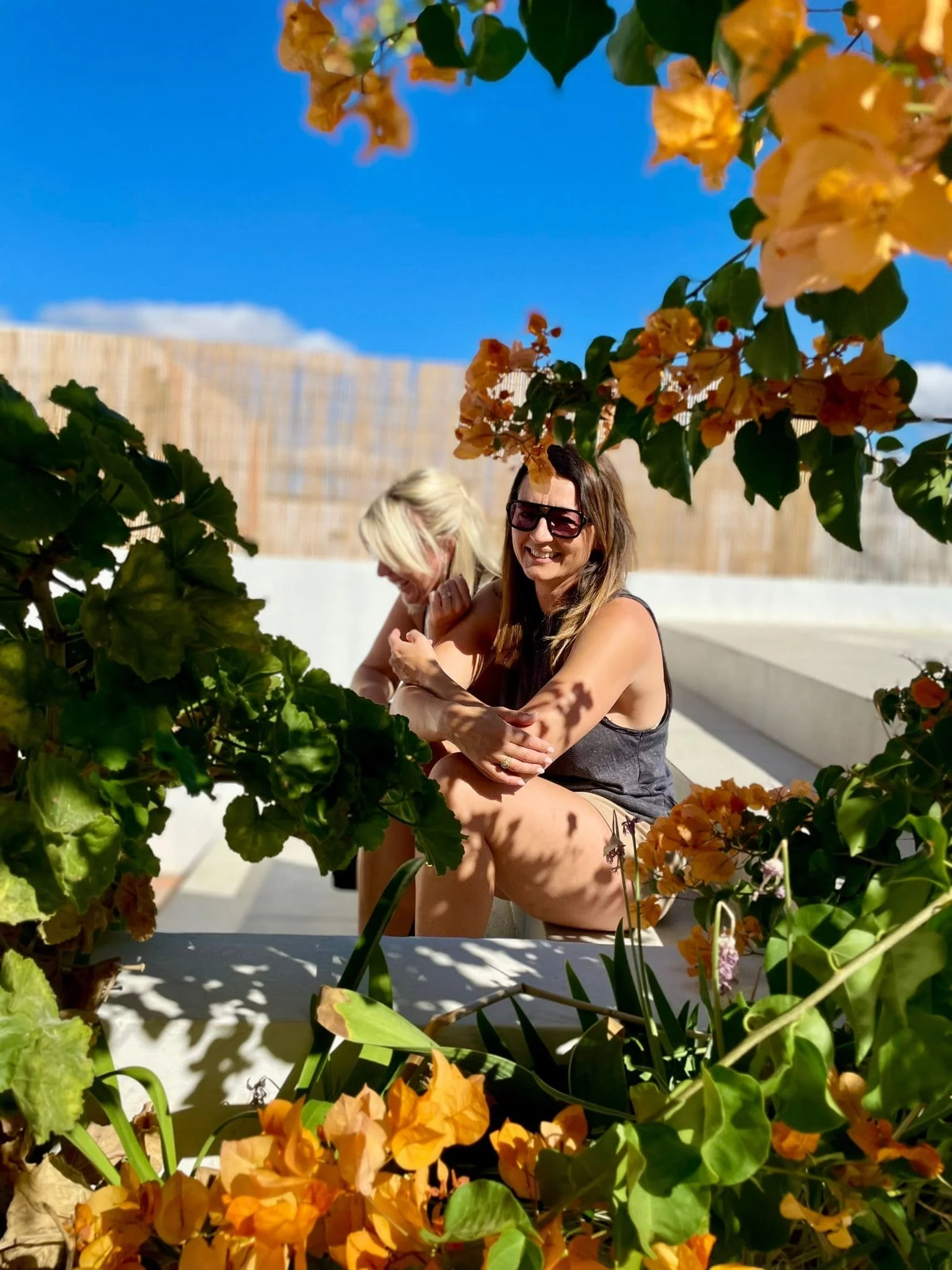 Two women sitting on a concrete bench under flowering plants with orange and pink blossoms, enjoying sunny weather with a bright blue sky and a bamboo fence in the background.
