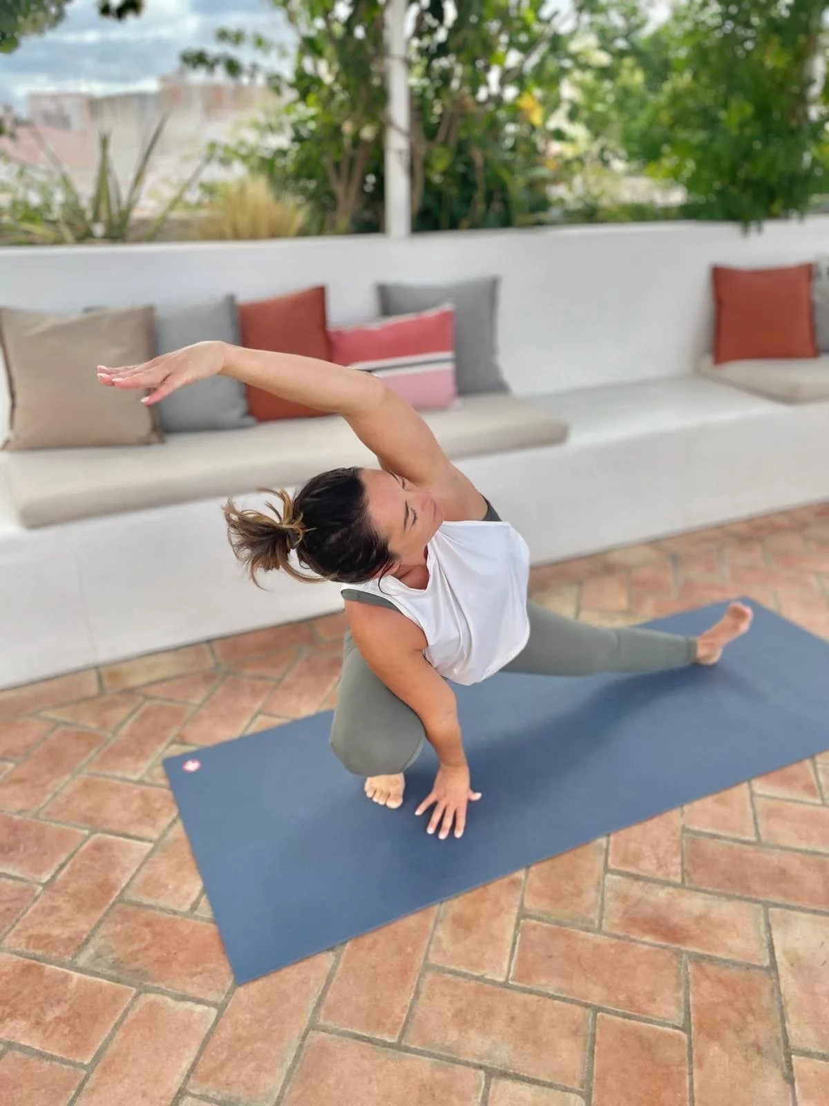 Woman practicing yoga outdoors on a blue mat. She is balancing in a one-legged pose with one arm extended overhead and the other hand on the ground. She is surrounded by a patio with terracotta tiles and a white bench with various colorful cushions i