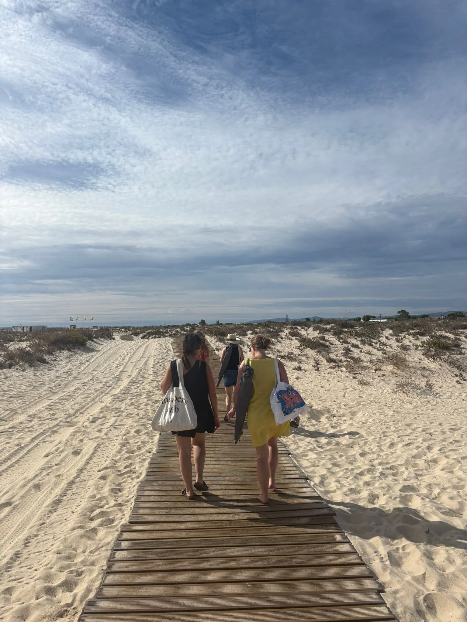 Four women walking on a wooden walkway towards the beach, carrying bags, with sand dunes and cloudy sky in the background.