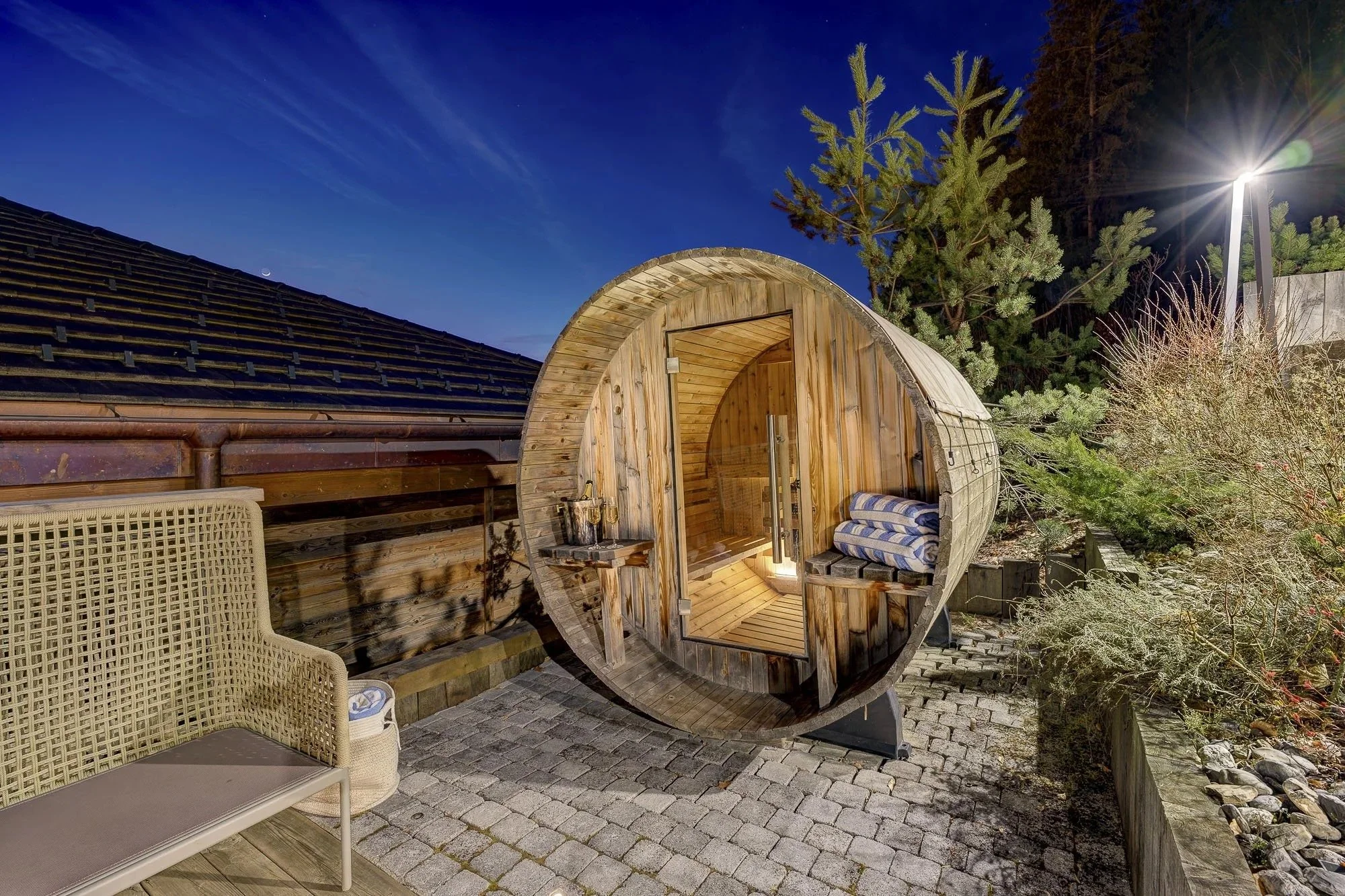An outdoor scene at night with a wooden barrel sauna, a wicker chair, a stone paved area, trees, and a bright streetlight illuminating the scene under a dark blue sky.