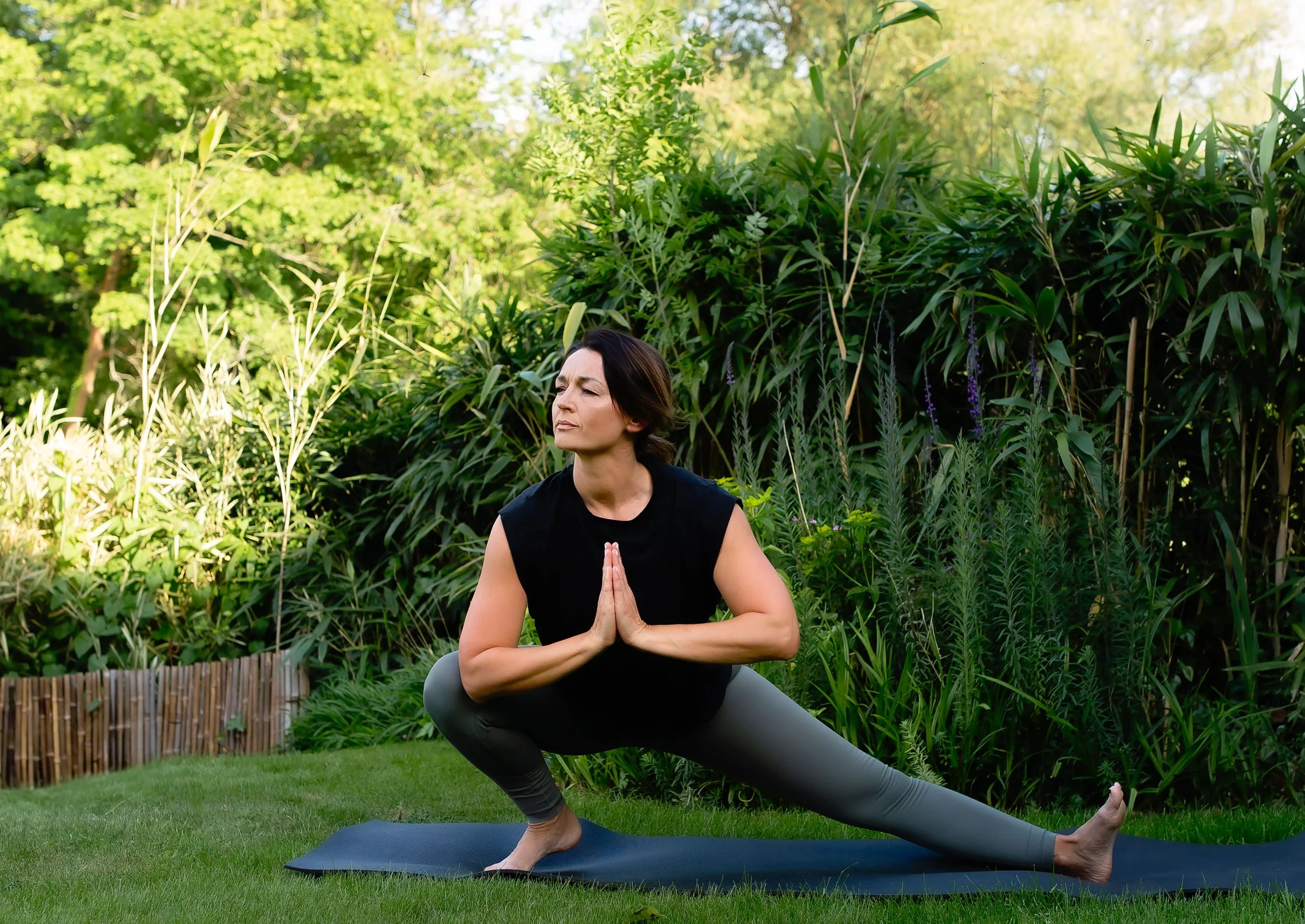 A woman practicing yoga outdoors on a mat in a garden, wearing a black sleeveless top and gray leggings, in a warrior pose with hands pressed together in front of her chest, surrounded by green foliage and trees.