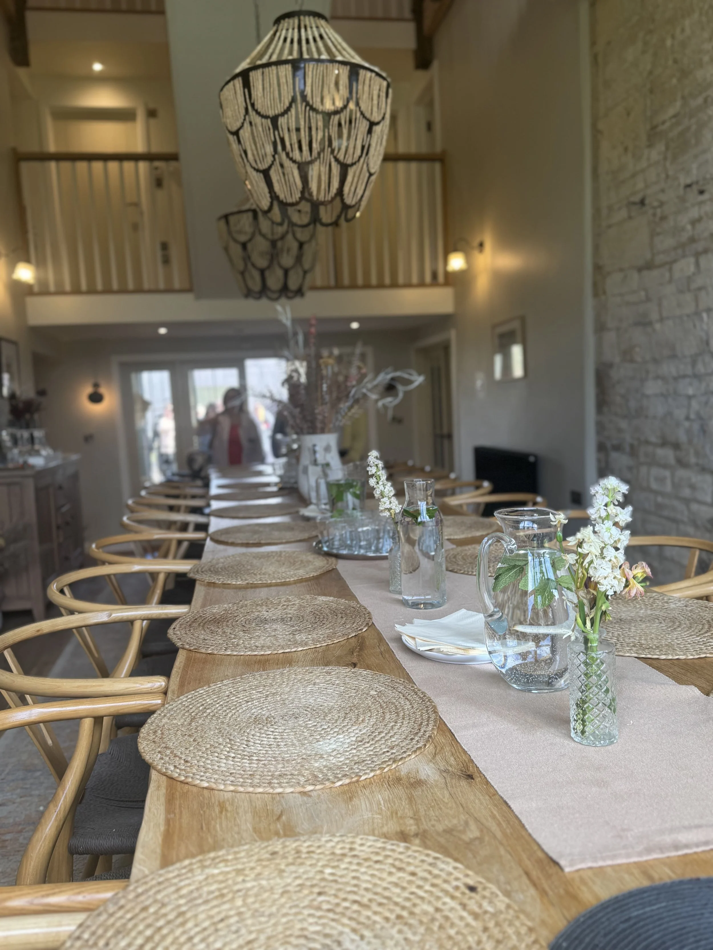 A long dining table set with woven placemats, glass pitchers, floral arrangements in vases, and napkins, with wooden chairs surrounding it in a cozy, well-lit interior featuring a chandelier, stone wall, and windows.