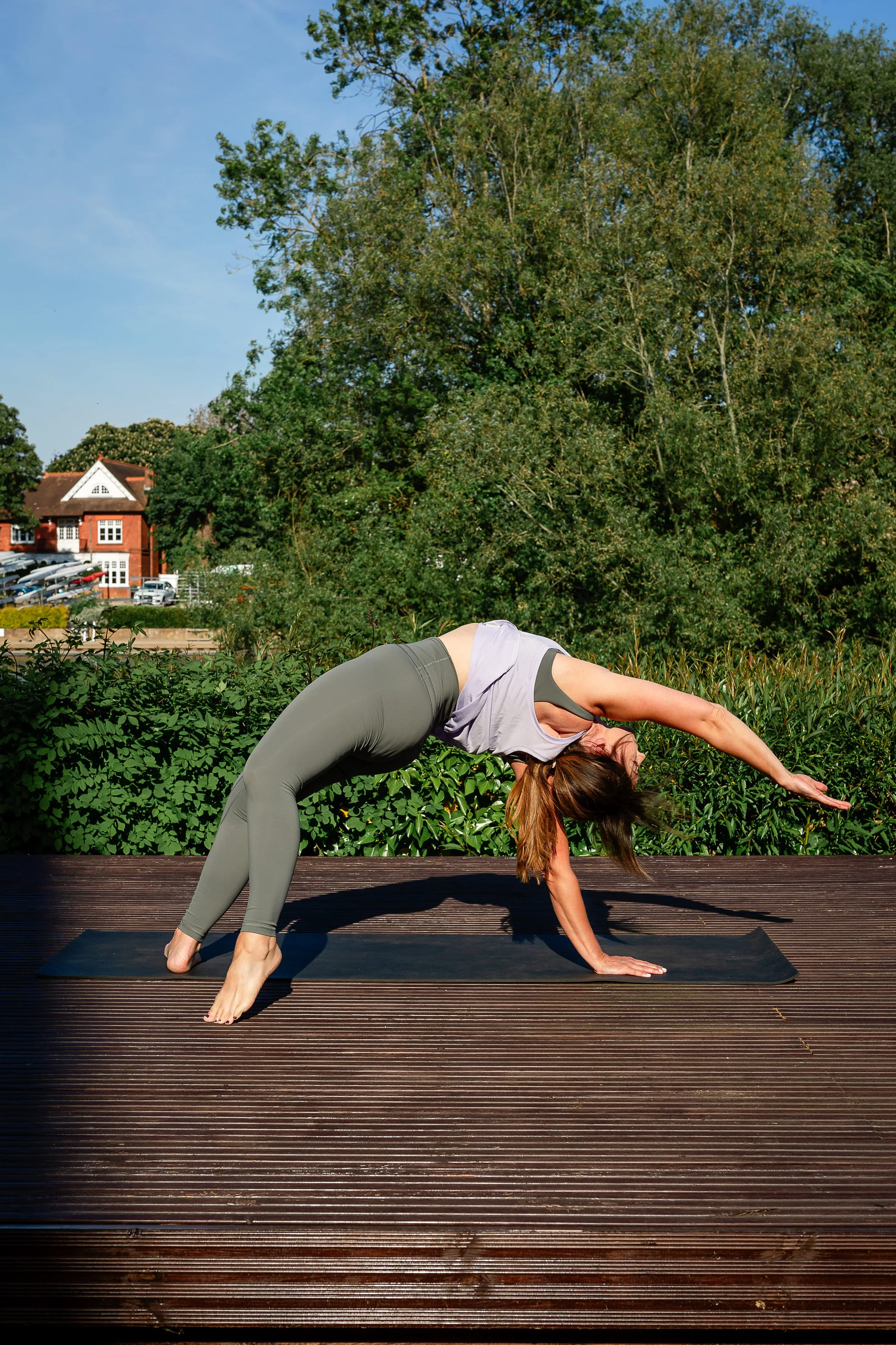 A woman practicing yoga outdoors on a wooden deck, performing a downward dog pose with her hands and feet on the ground and hips raised. She is wearing a gray tank top and gray leggings, with a backdrop of lush green trees and a building in the distance under a clear blue sky.