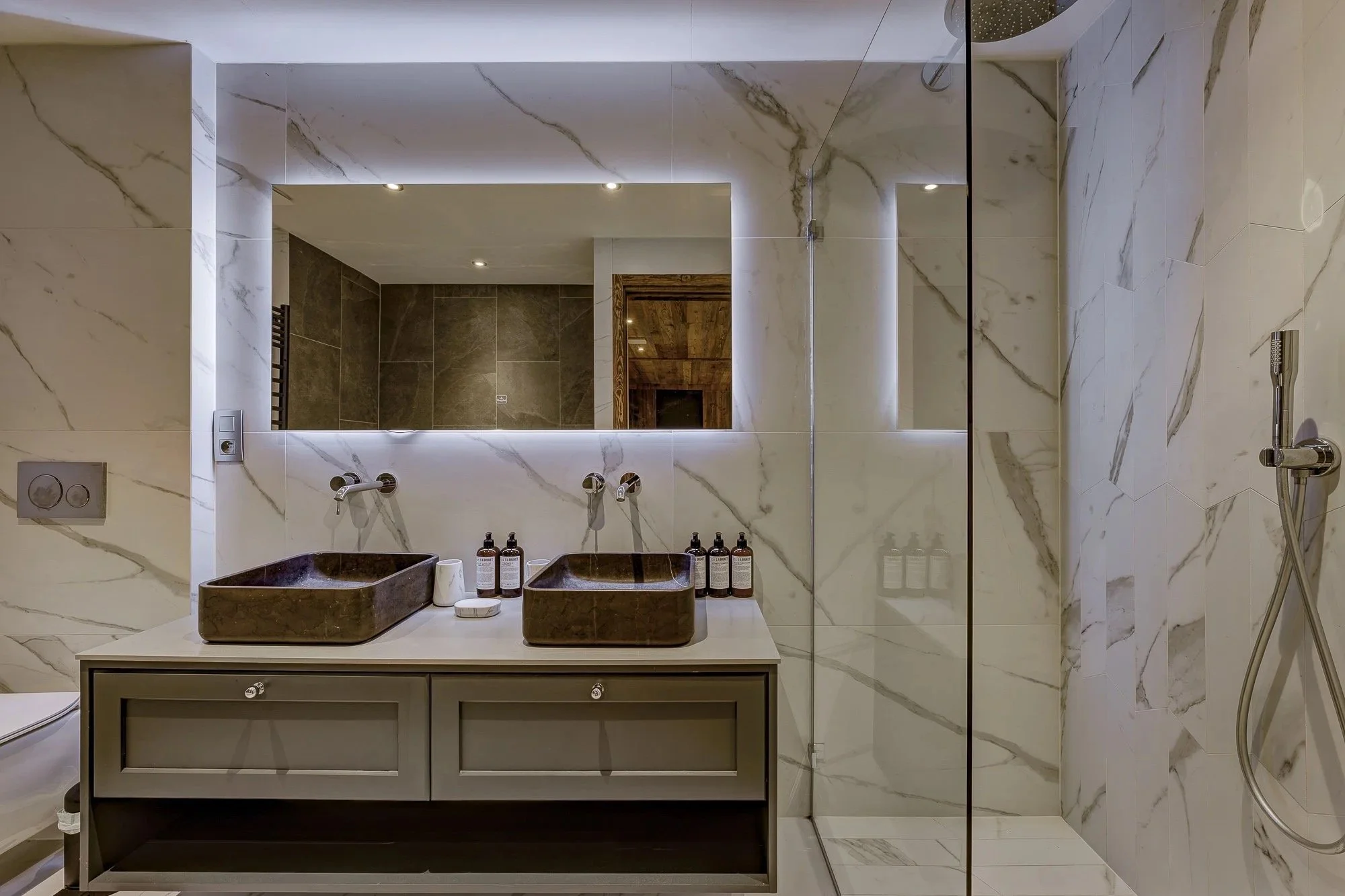 Modern bathroom with marble walls and dual brown sinks on a beige vanity, large mirror with backlighting, and a shower area with a handheld showerhead, bottles of toiletries on the counter, and a reflection of wooden ceiling panels.